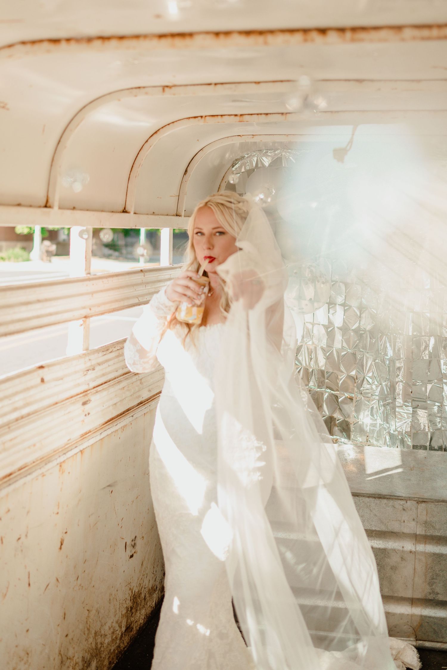 A bride in white dress poses inside a vintage bus with sunlight streaming through windows.