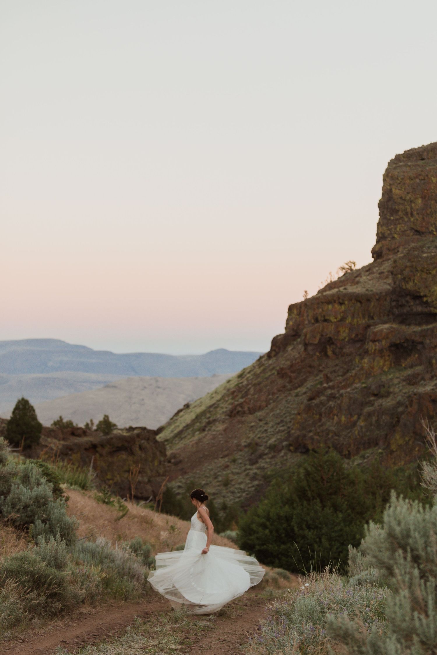 A white wedding dress twirls against a dramatic mountain landscape at sunset.