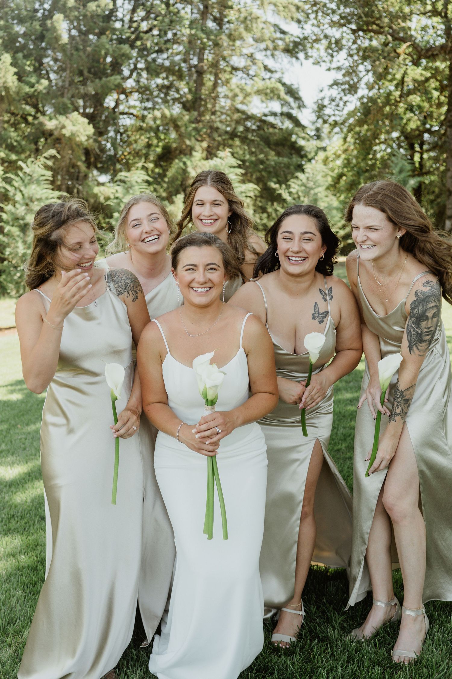 A group in champagne-colored dresses sharing a candid laughing moment on a sunny lawn.