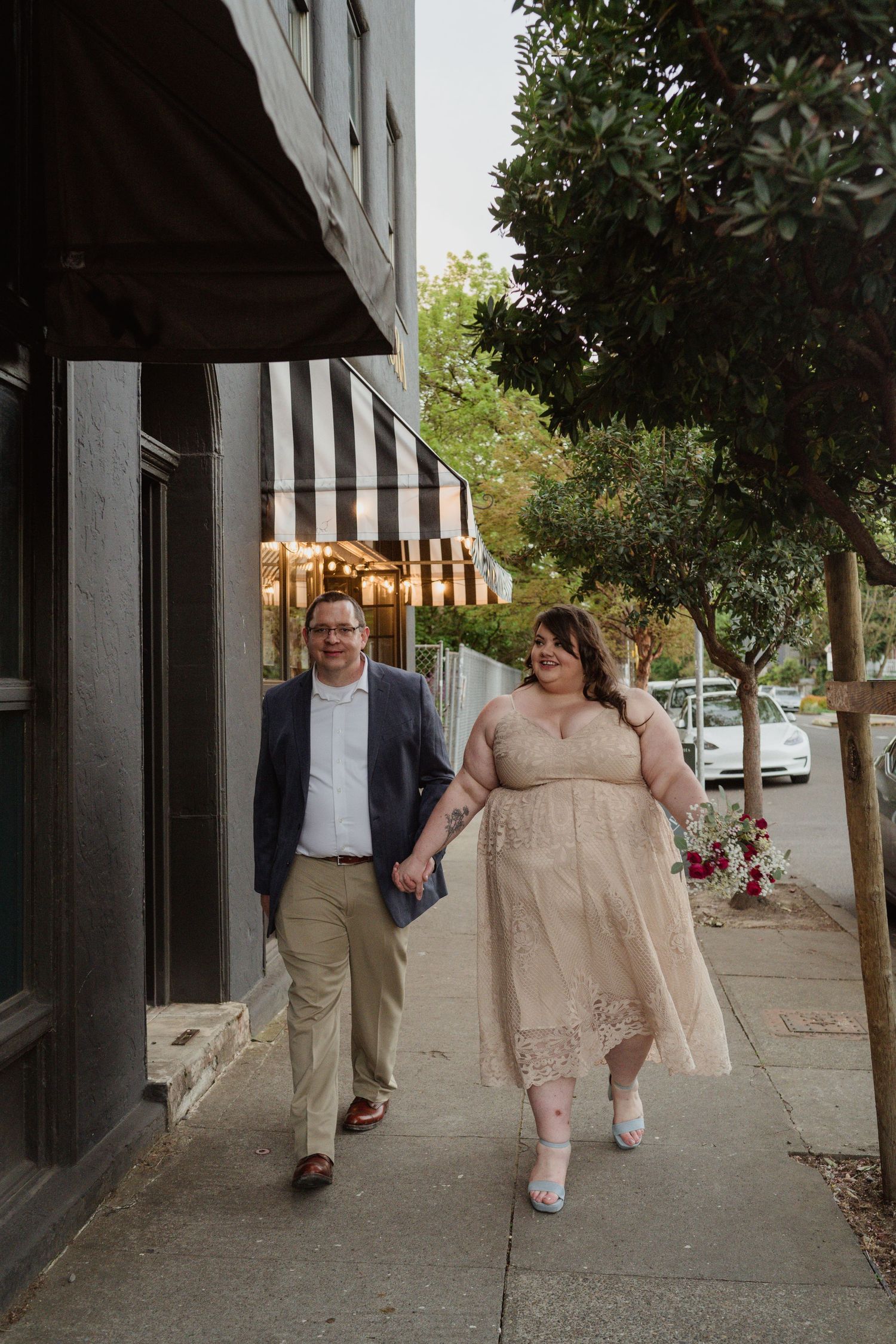 A couple walks hand in hand along a sidewalk with striped awnings and trees.