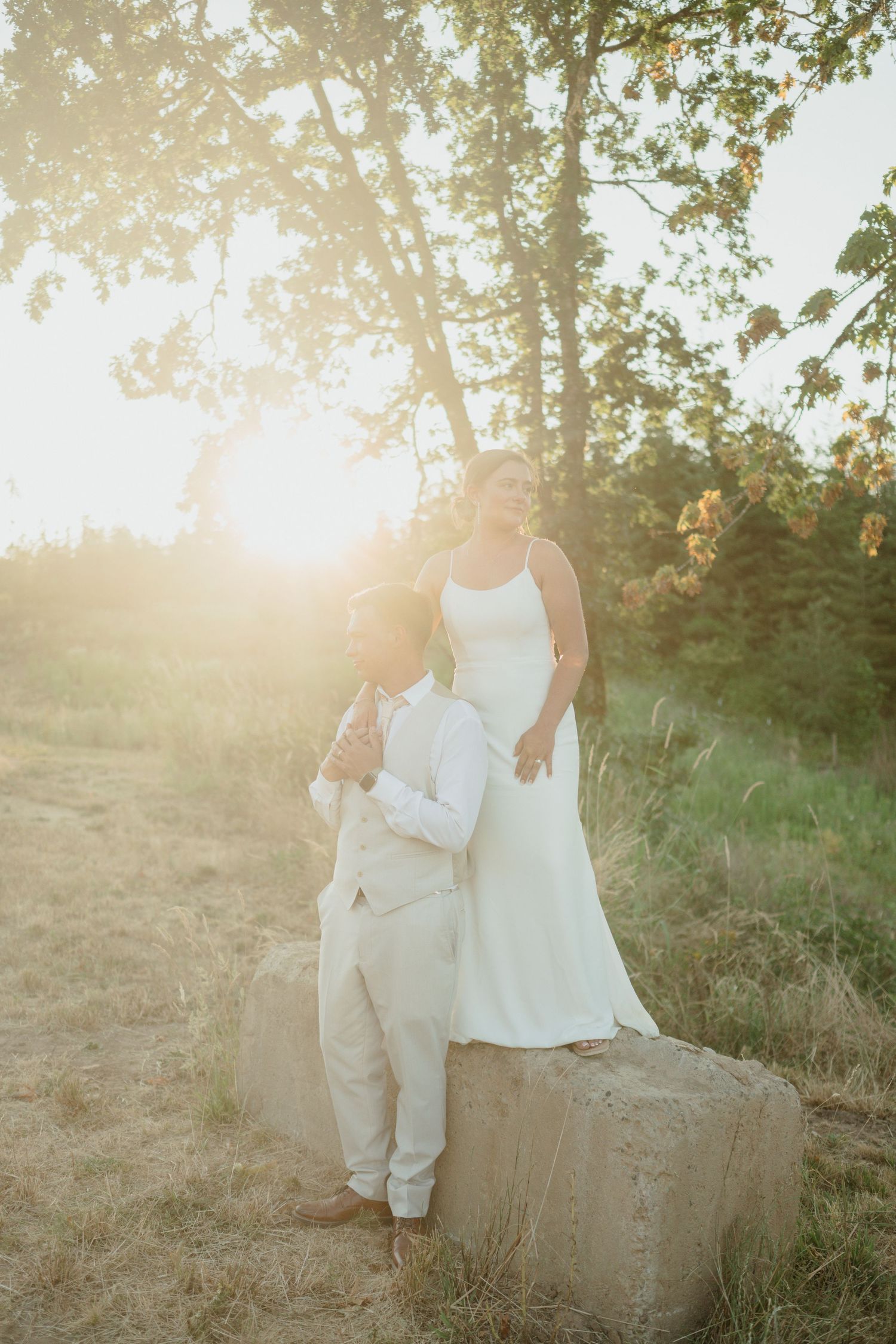 A romantic sunset portrait of a couple in white wedding attire standing on a rock.