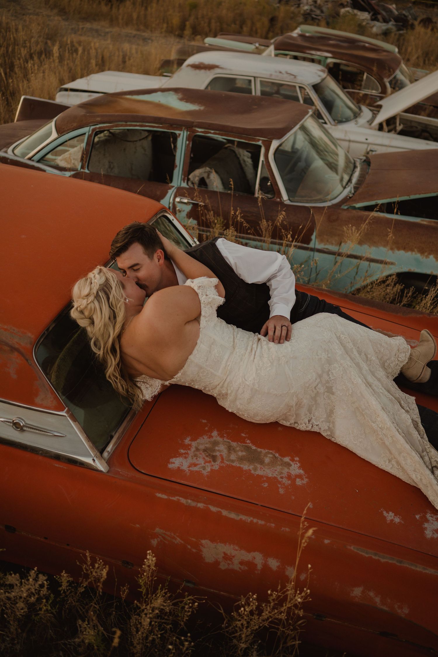 Newlyweds share intimate moment on vintage red car in rustic junkyard setting.