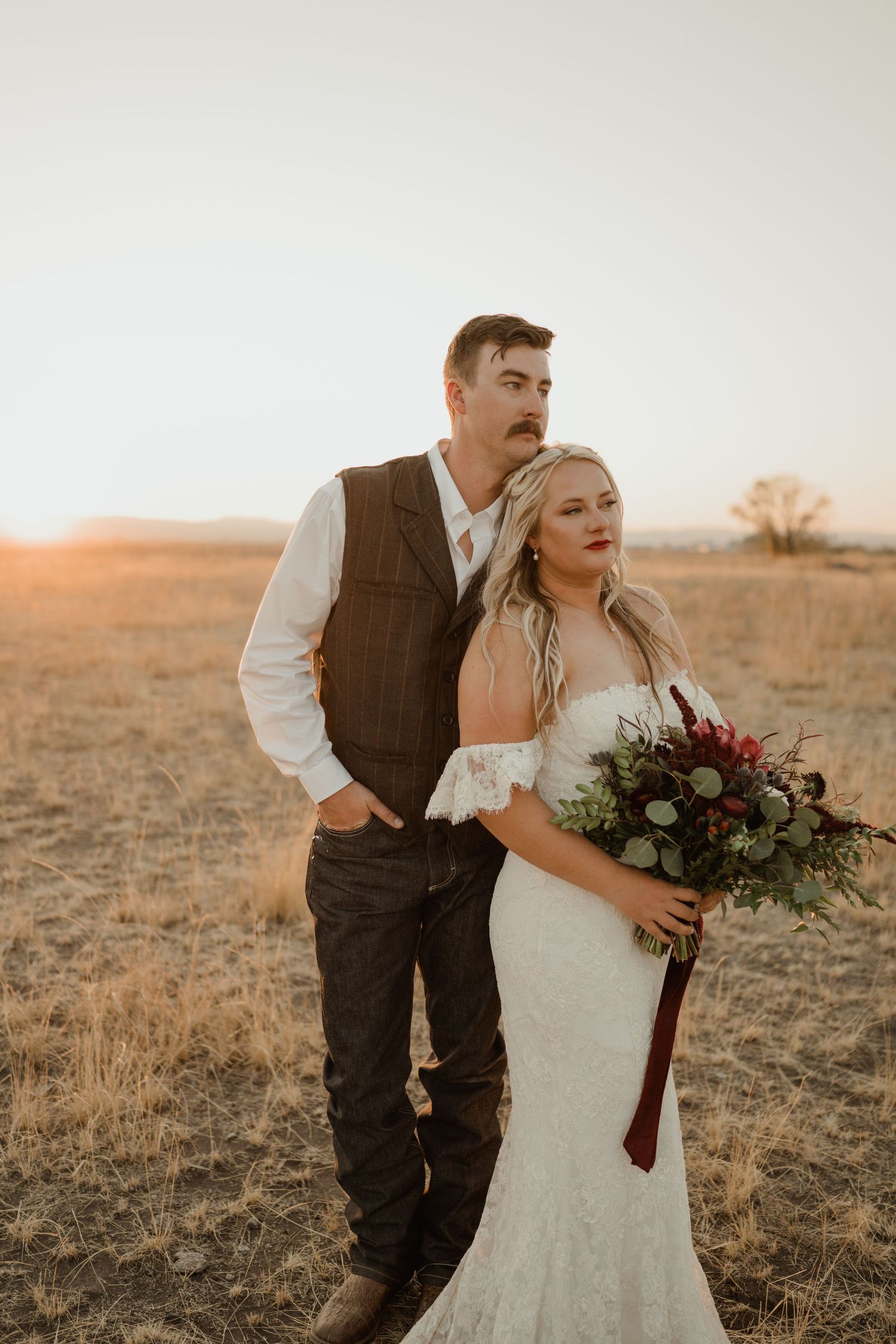 A rustic wedding portrait of a couple in a field at golden hour with a burgundy bouquet.