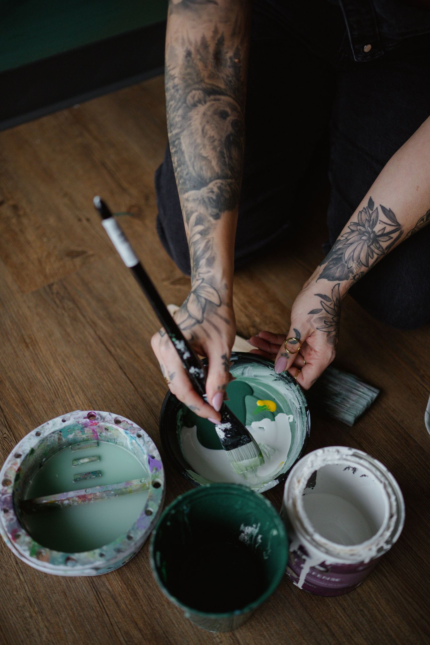 Tattooed hands mixing green paint in various containers on a wooden surface.