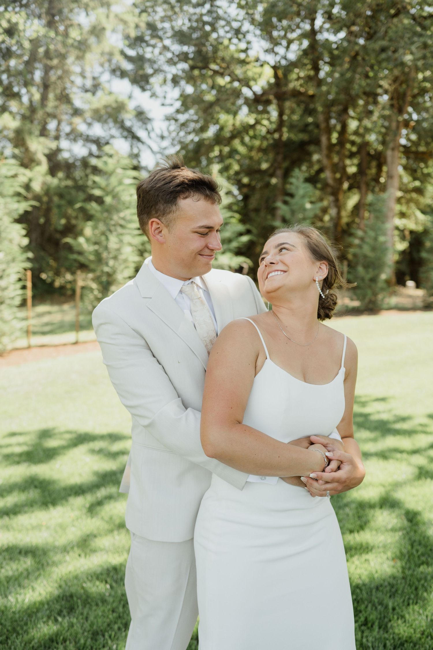 A tender embrace between newlyweds in white attire on a sunny lawn.