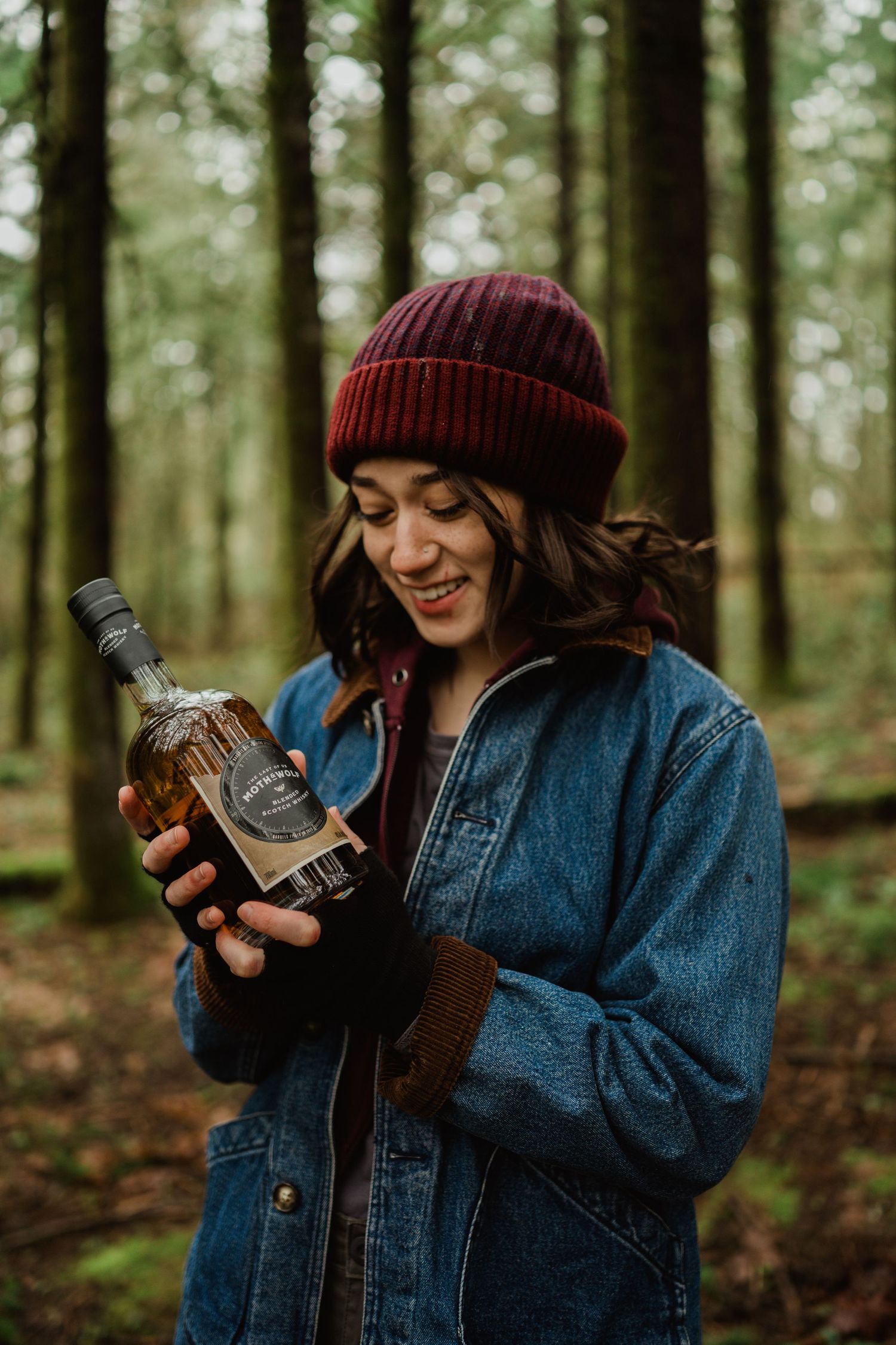 A person in a denim jacket and red beanie holds a bottle of whiskey in a forest setting.