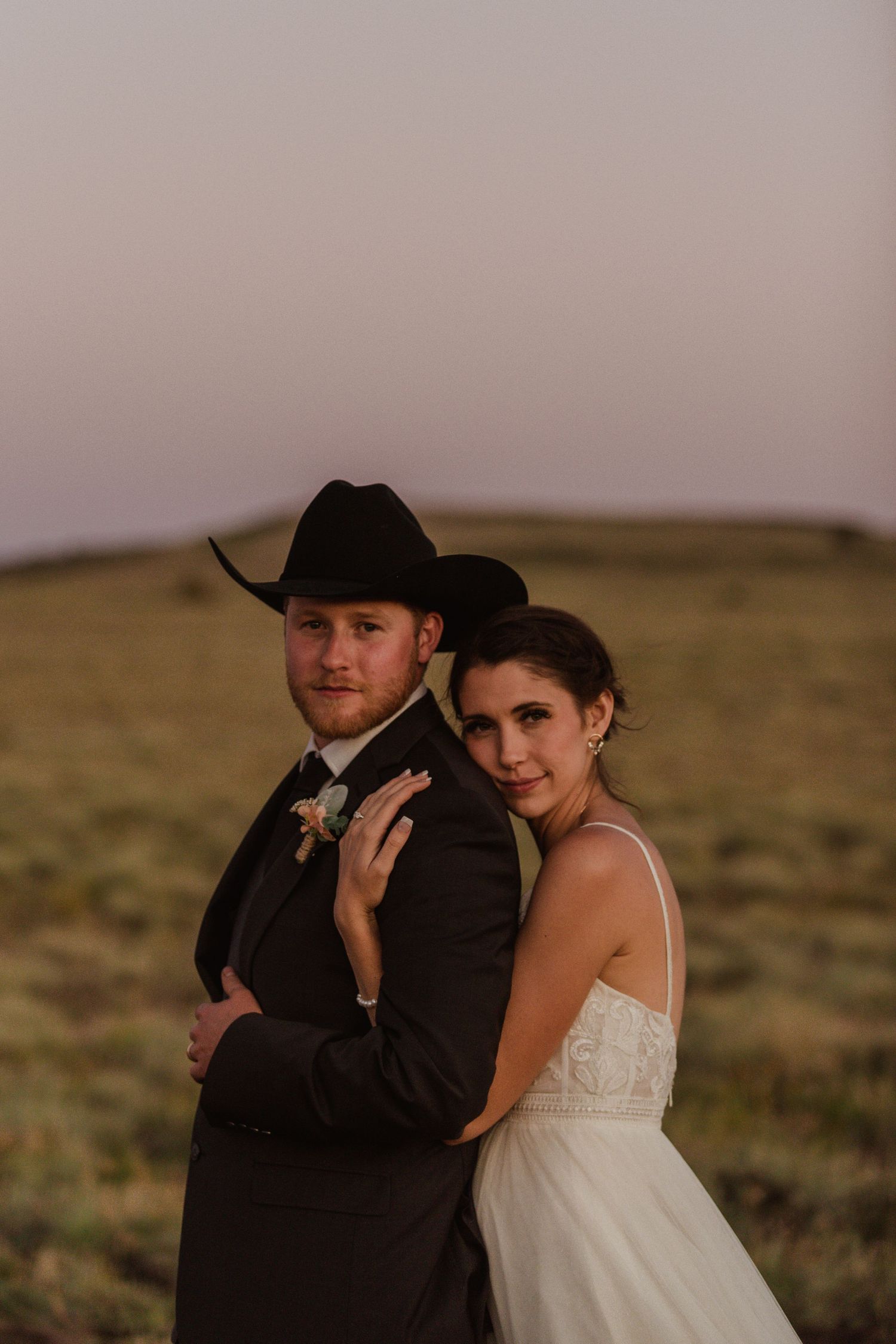 A western-themed wedding portrait shows a couple embracing on a hillside at dusk.