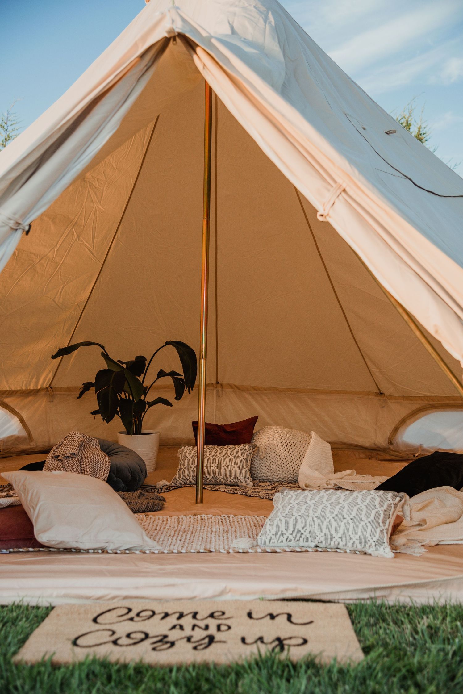 Interior of a bell tent decorated with cozy pillows and blankets, with a welcome mat at entrance.