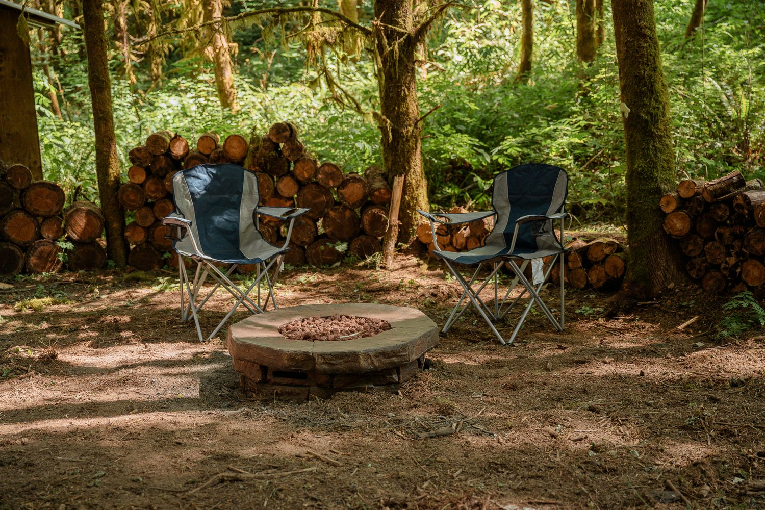 Outdoor camping setup with blue chairs around stone fire pit in wooded area.