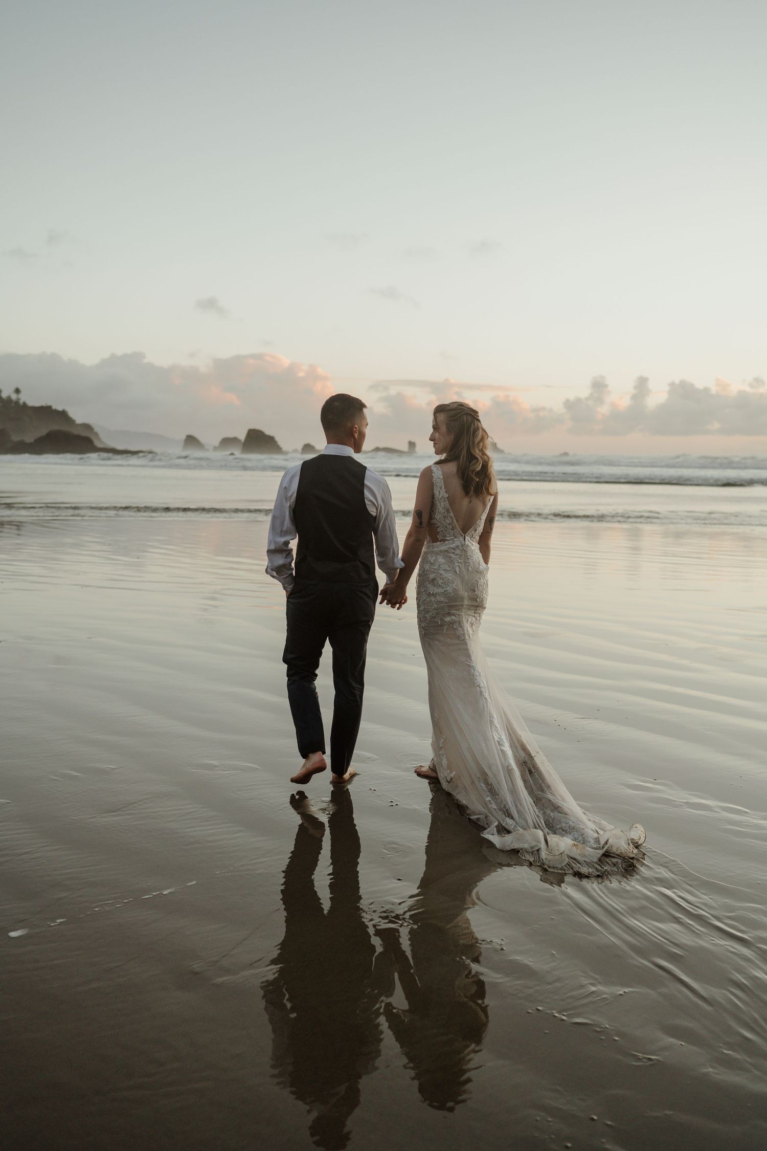 A romantic beach wedding photo with couple walking along reflective wet sand at sunset.