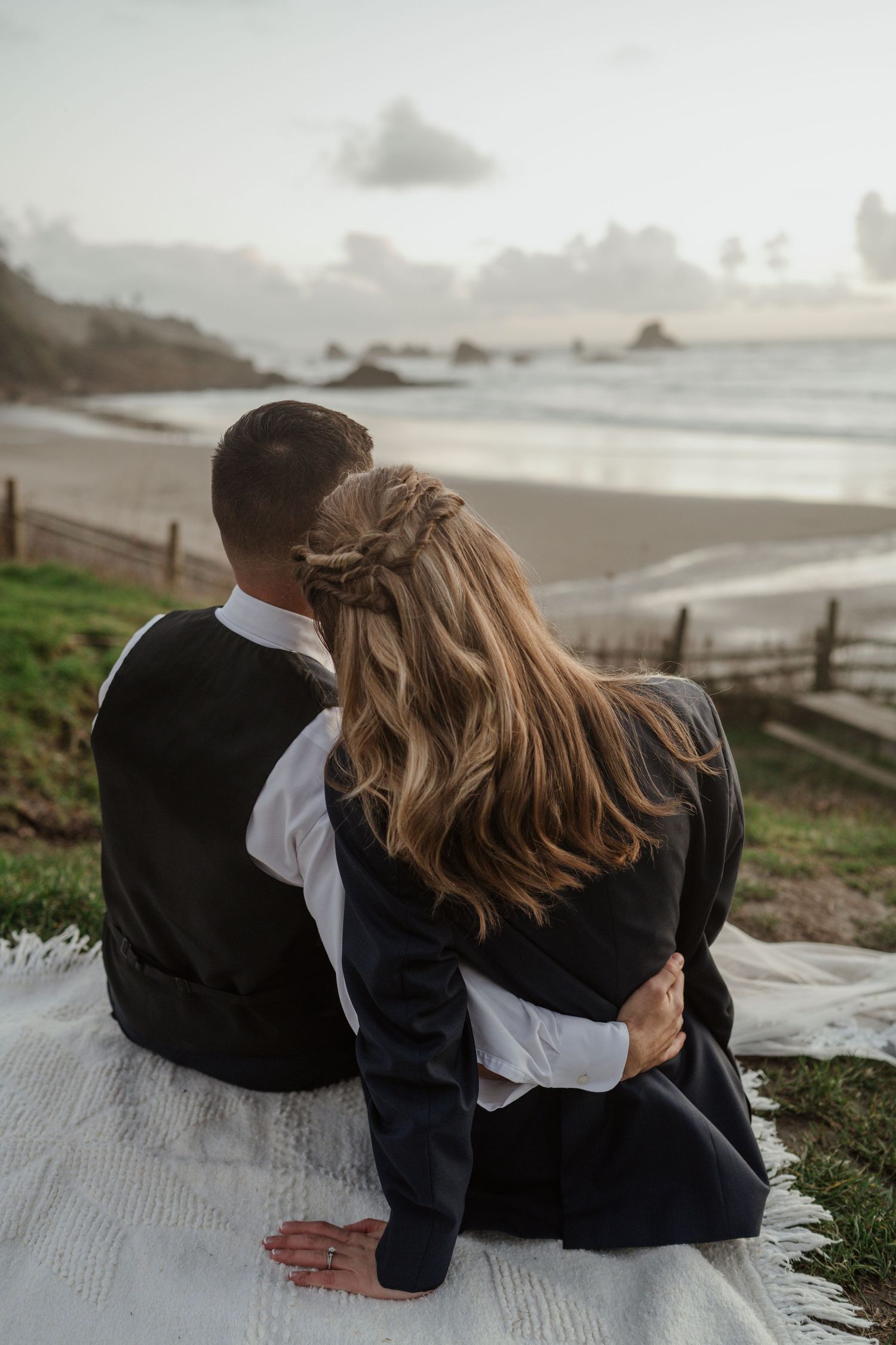 A couple sits together on a blanket overlooking a scenic coastal view at sunset.