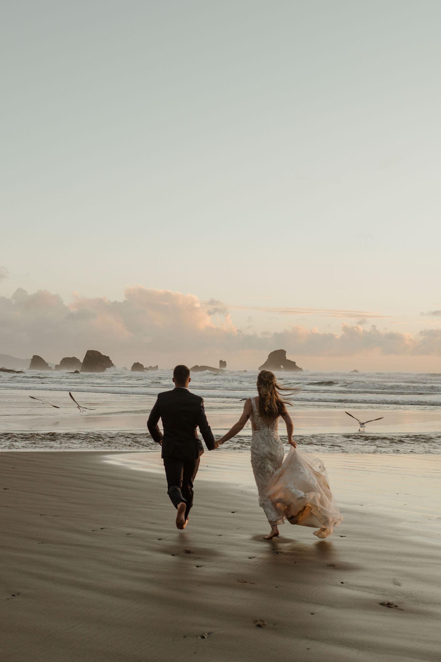 A couple runs joyfully along a beach at sunset holding hands with seagulls flying overhead.