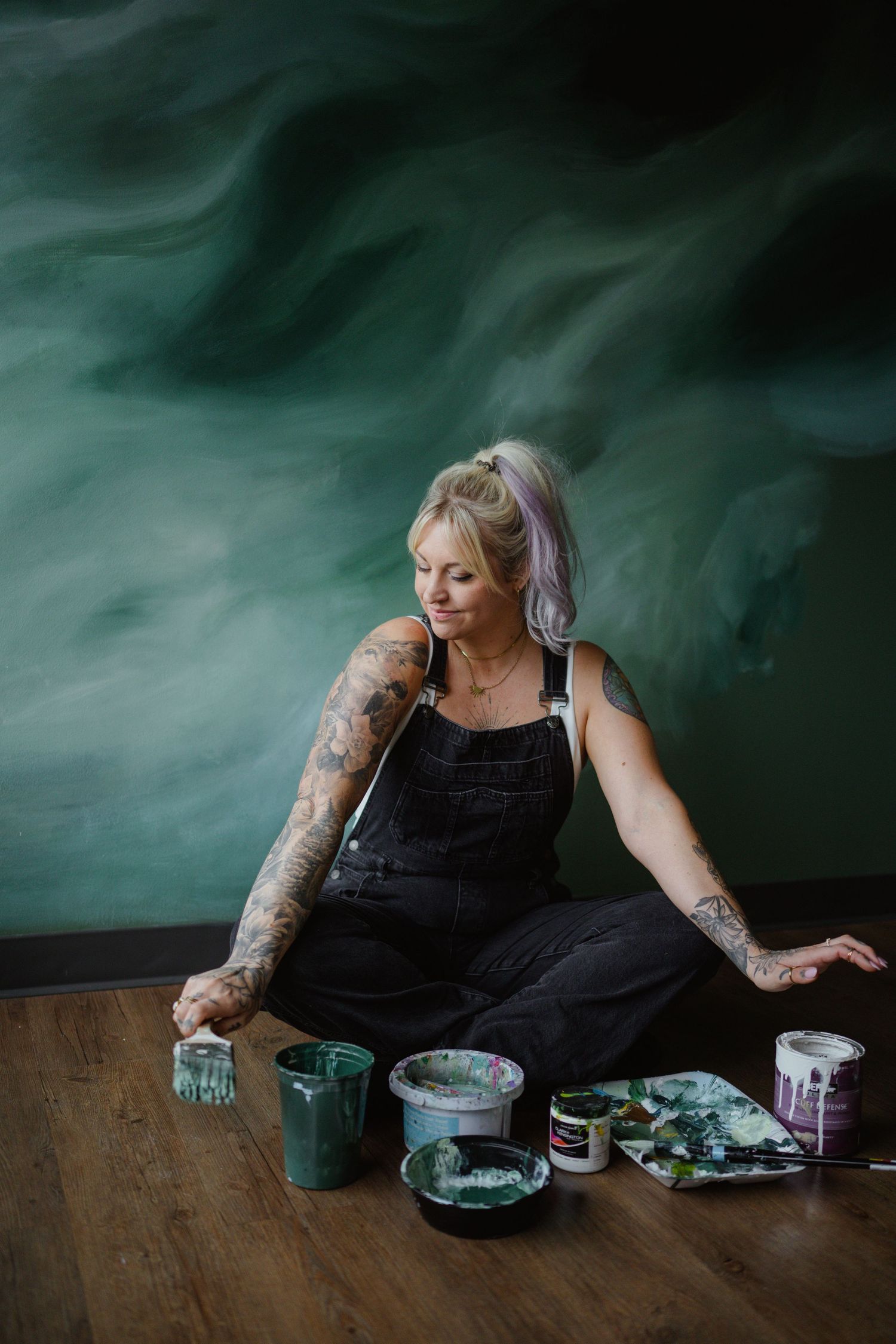 An artist sits with paint supplies against a moody green backdrop.
