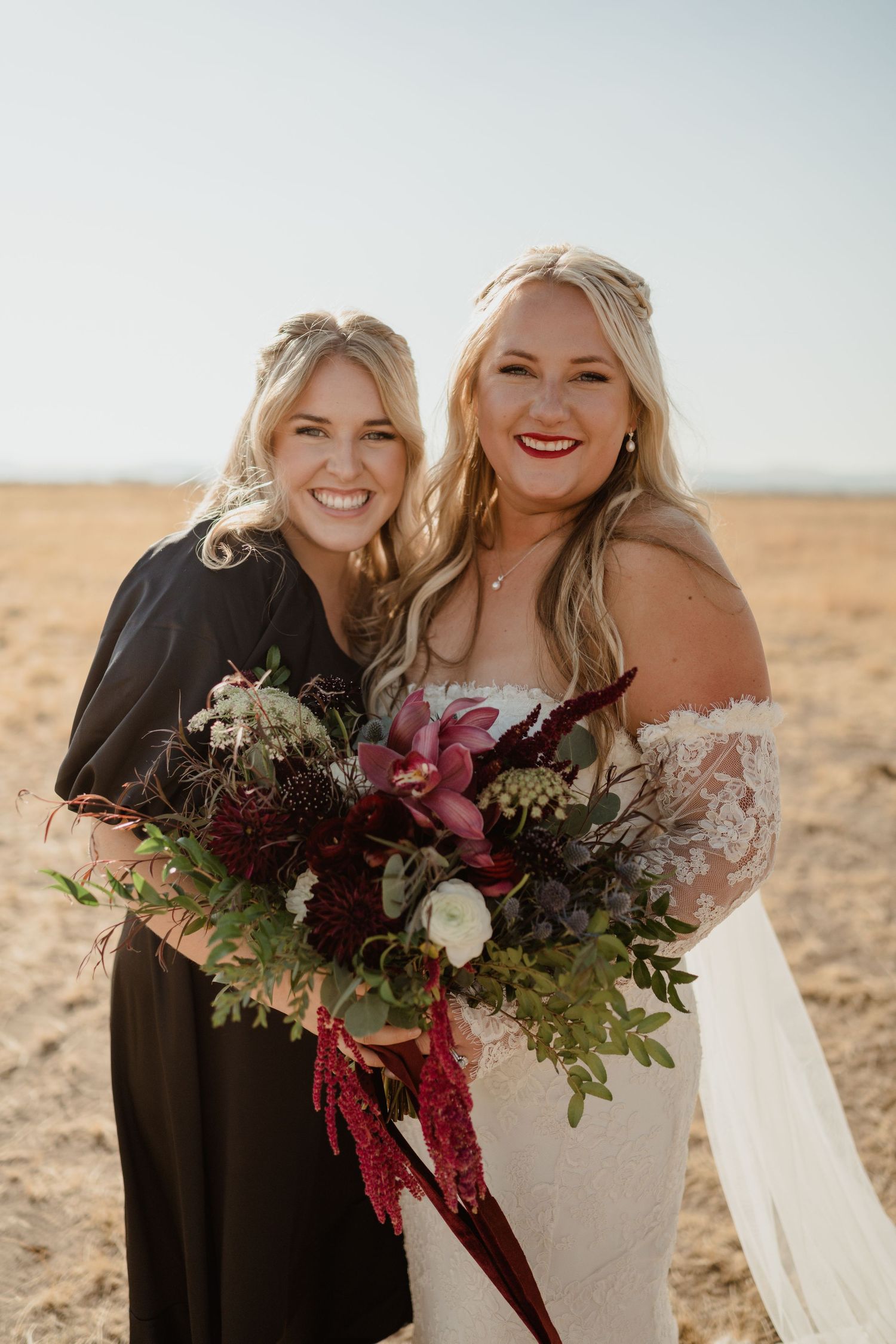 Two women pose with a burgundy and green wedding bouquet against golden field at sunset.