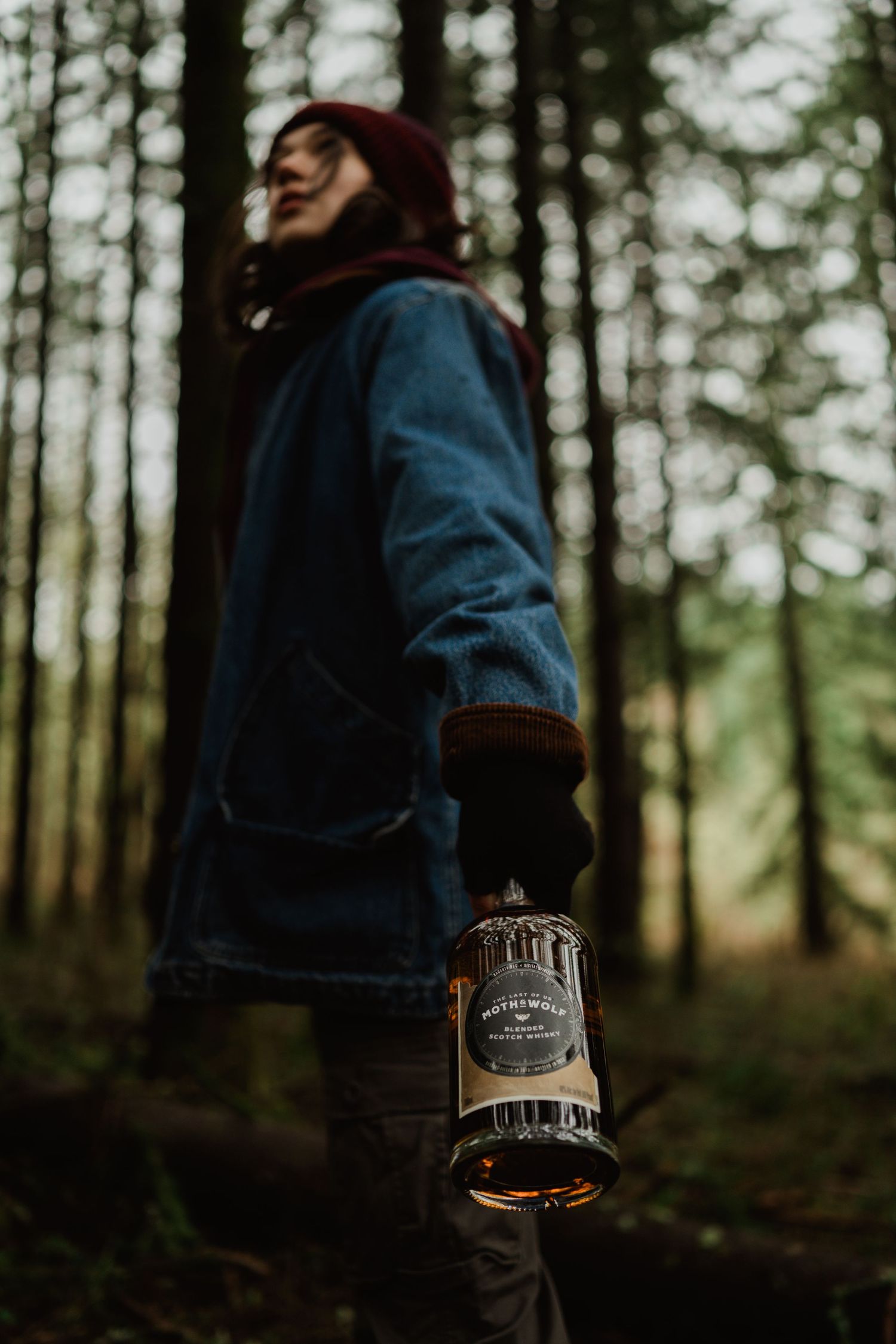 Someone wearing a denim jacket and red beanie holds a whiskey bottle in a misty forest.