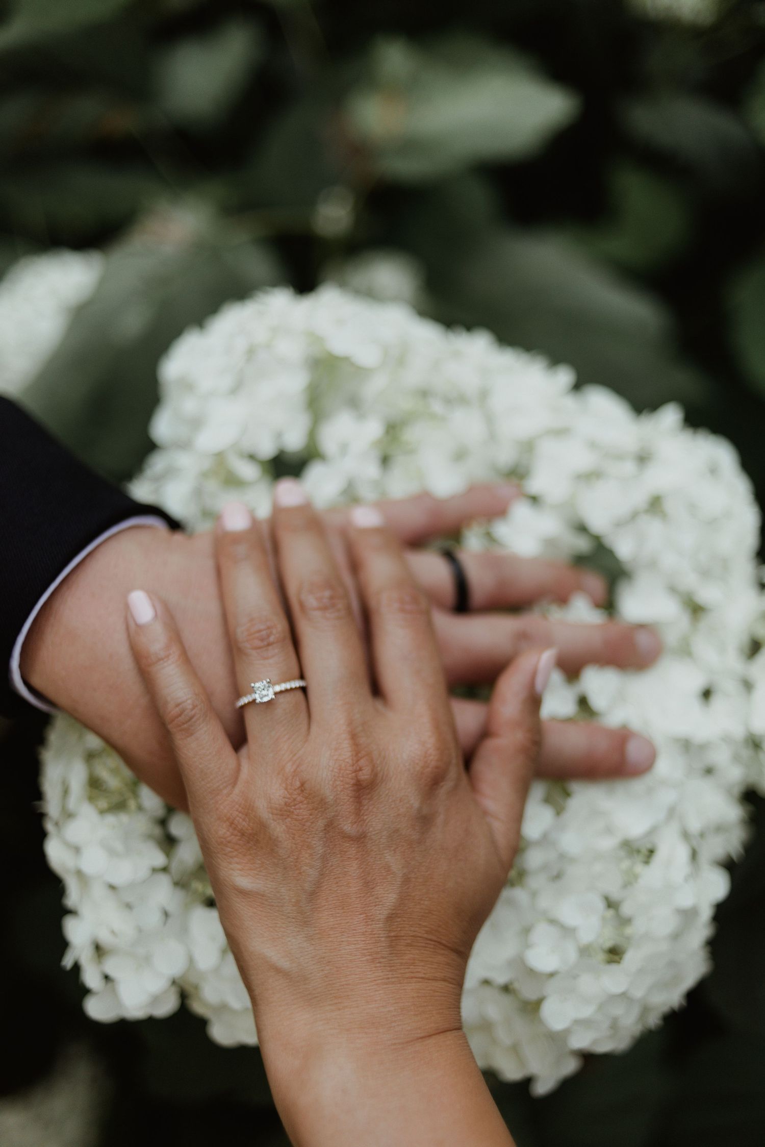 Close-up of wedding rings and interlocked hands against white hydrangea flowers.
