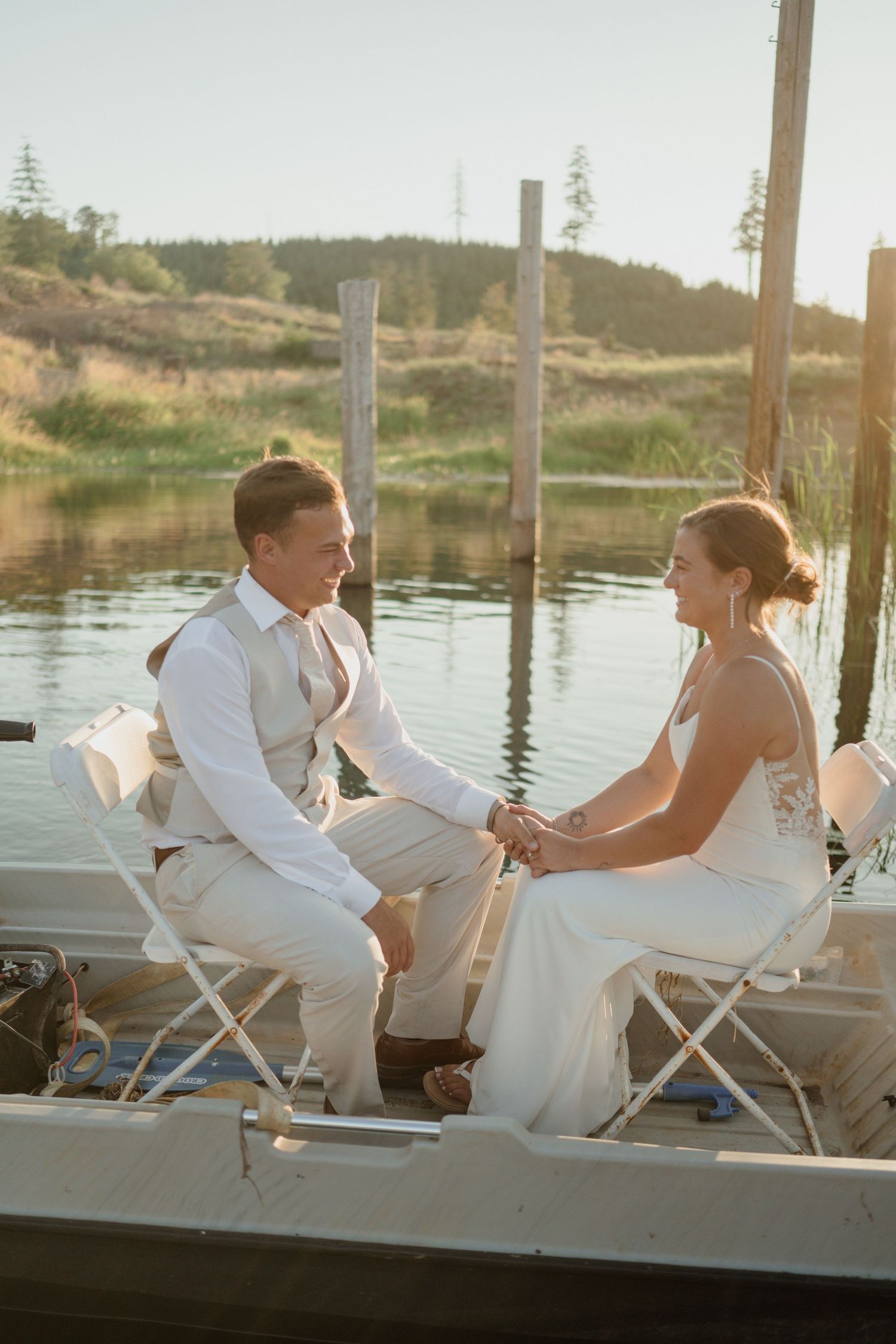 Newlyweds share an intimate moment while sitting on chairs on a dock by the water.