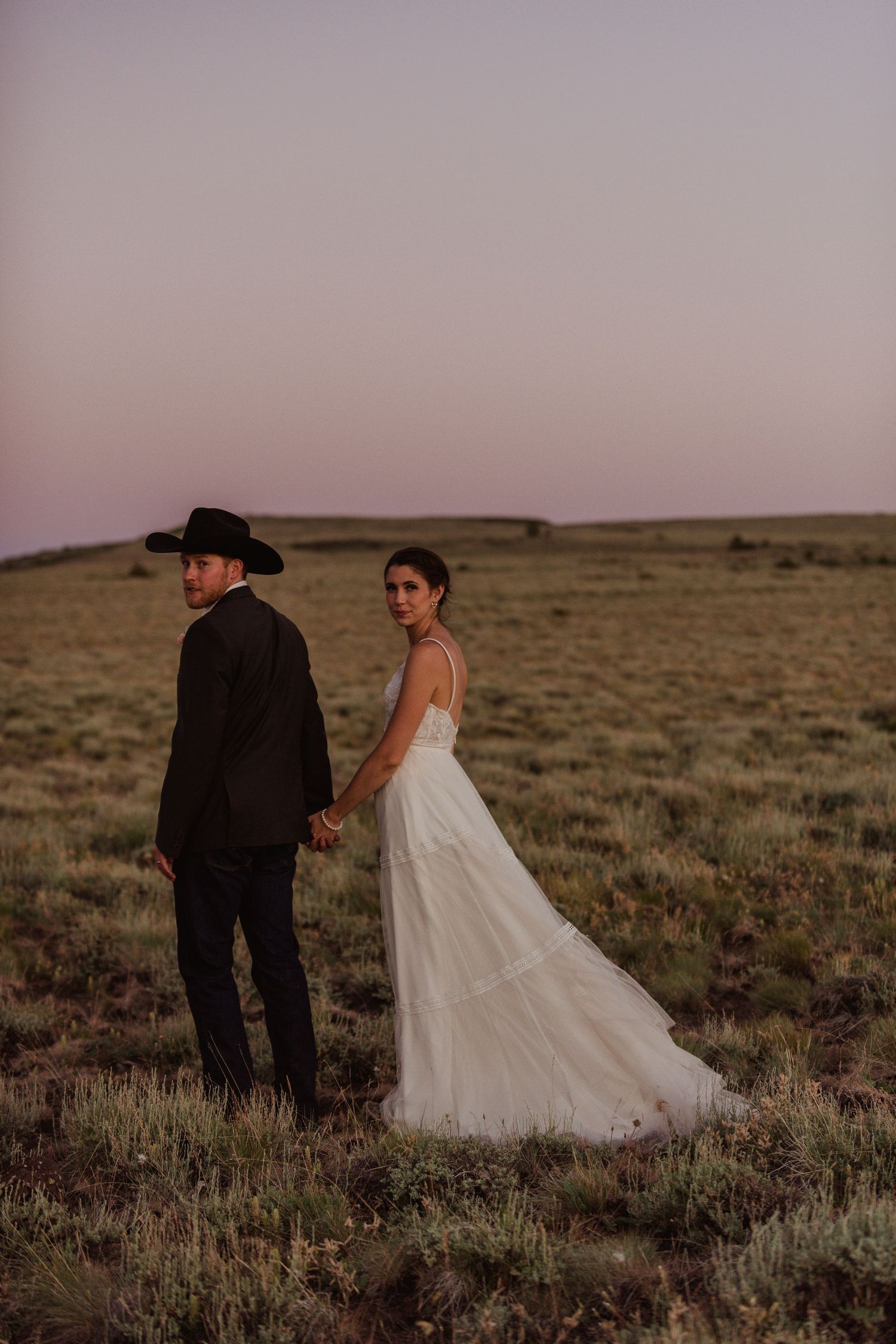 A couple in wedding attire stand hand-in-hand in a desert landscape at sunset.