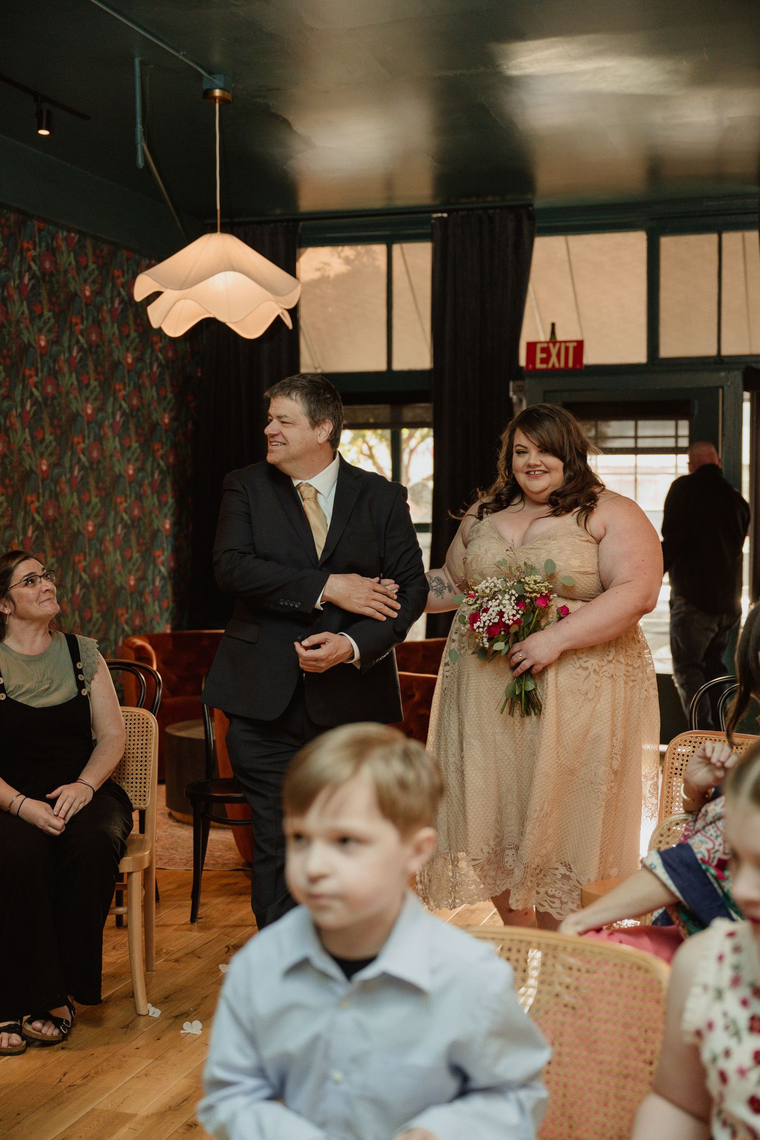 A couple enters a venue with floral wallpaper and modern lighting while guests look on during a wedding ceremony.