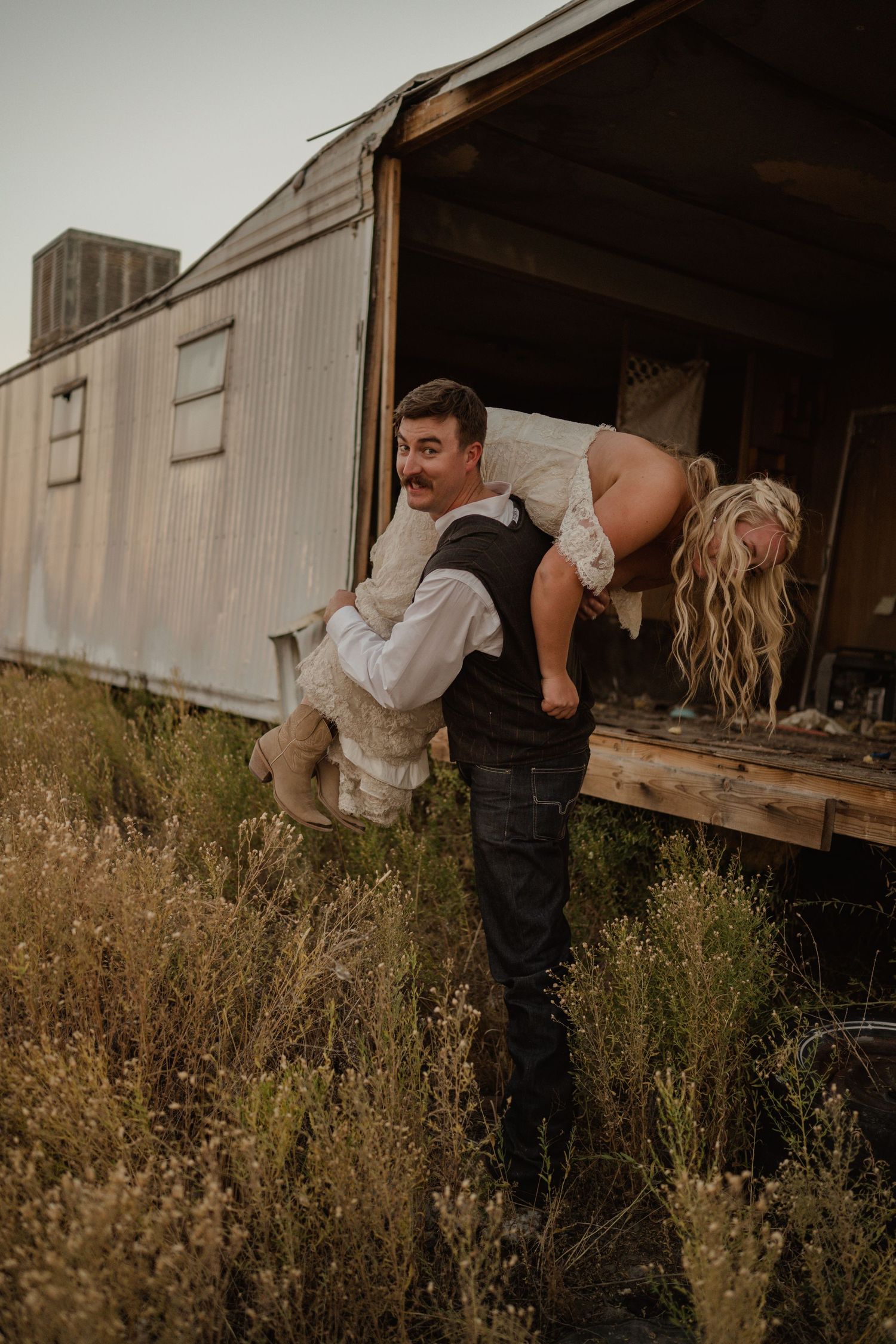 Playful wedding photo of a couple at an old train car surrounded by tall grass.