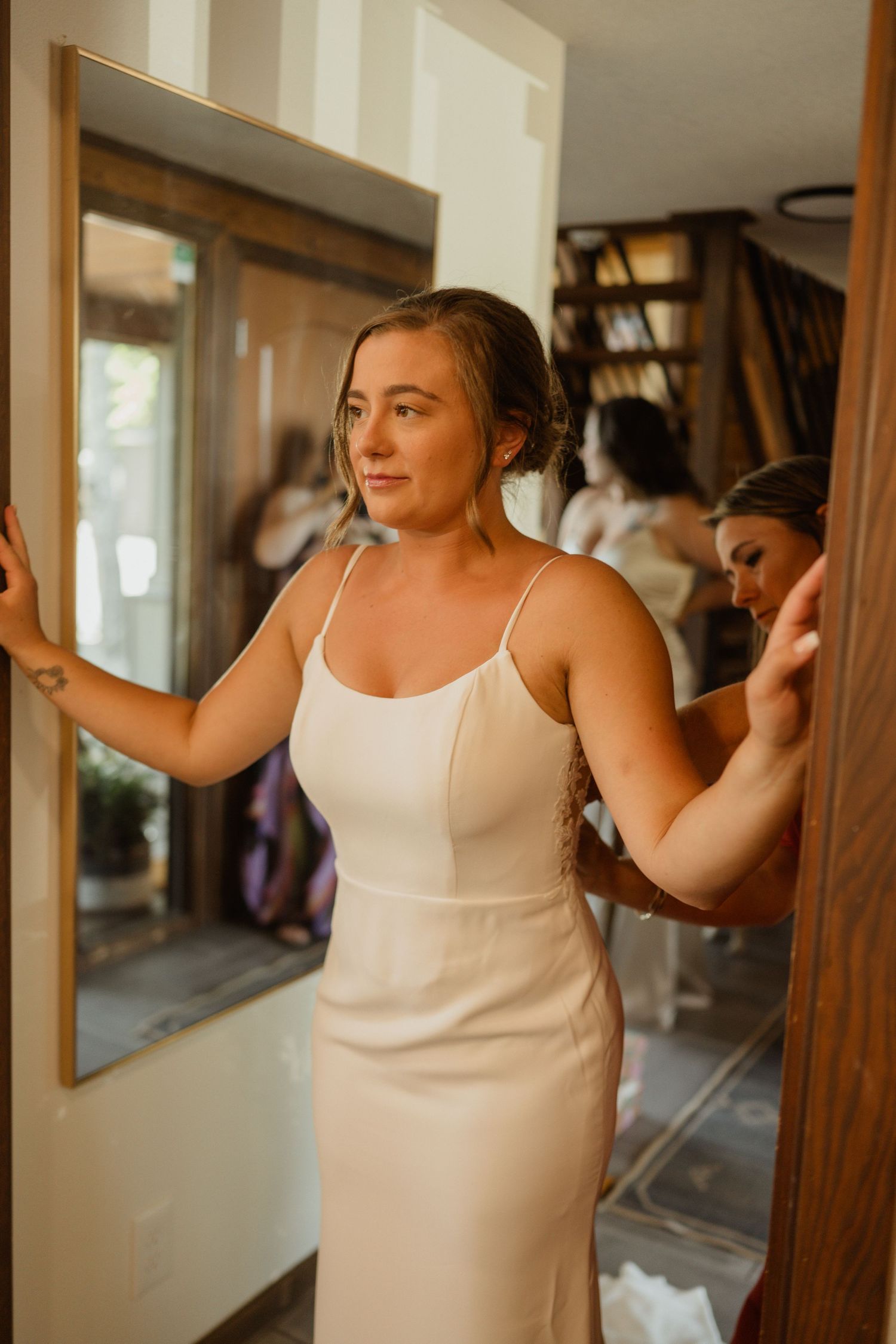 A person in a white spaghetti strap dress stands near a window in warm interior lighting.