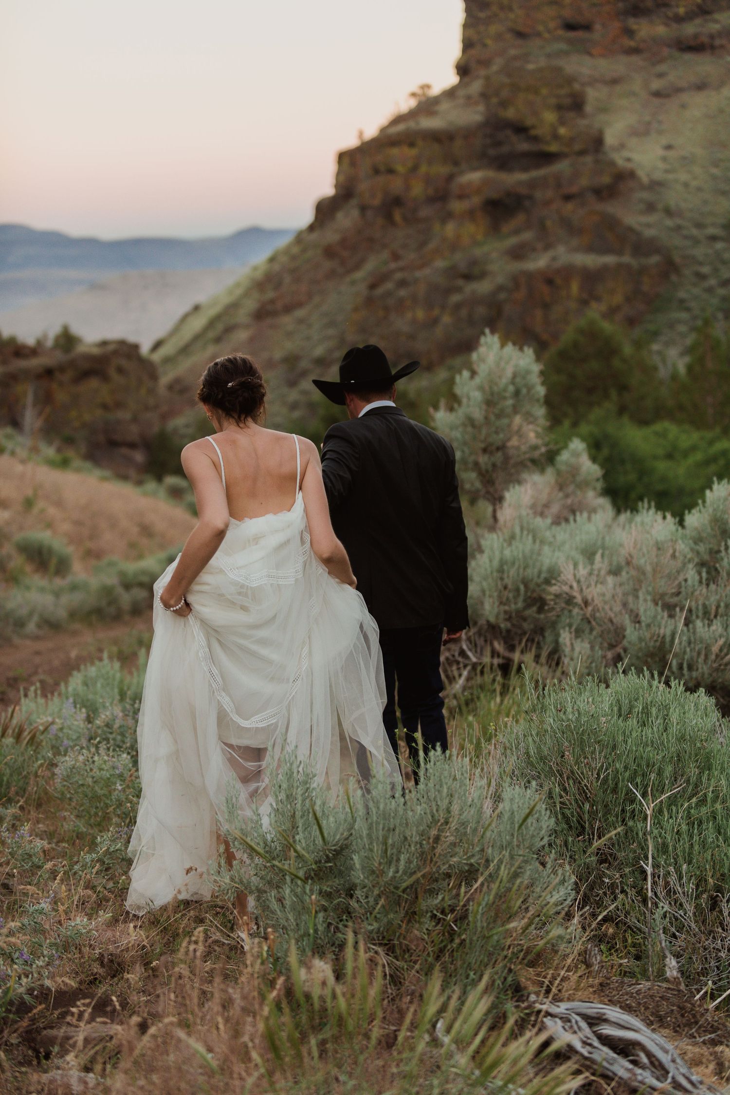 A couple walks through desert sagebrush with dramatic rocky cliffs in the background.