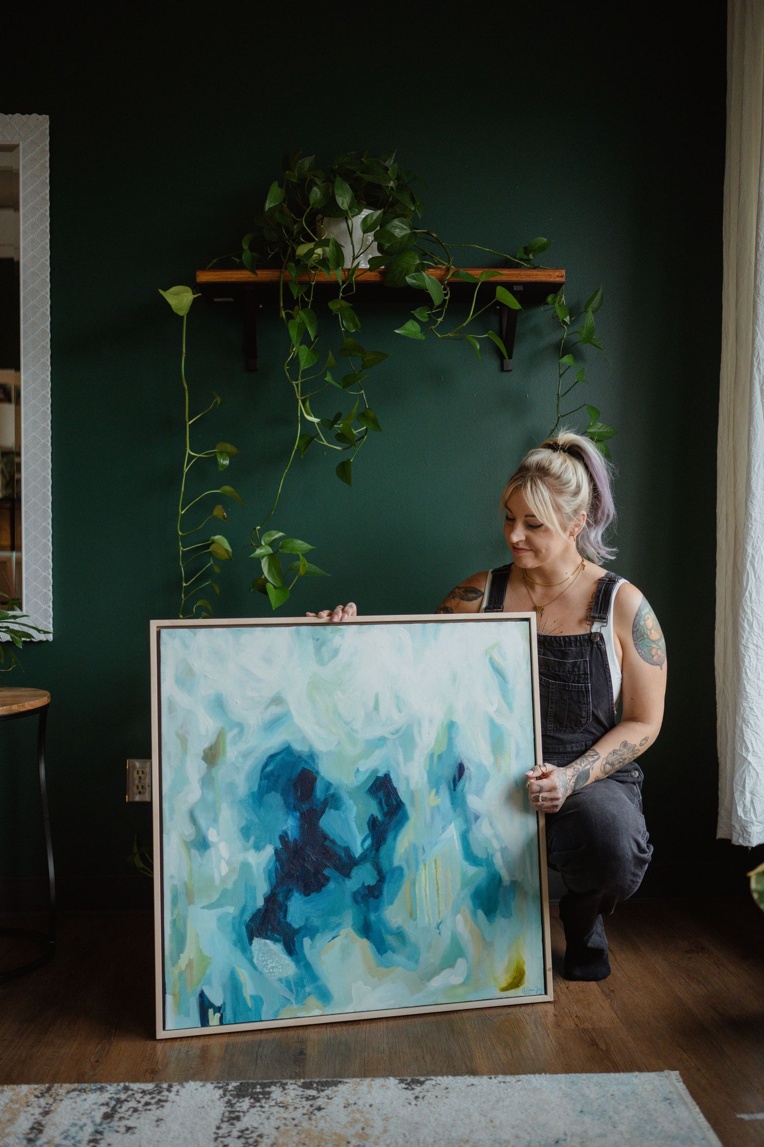 Artist showcasing a blue and white abstract painting in front of a green wall with hanging plants.