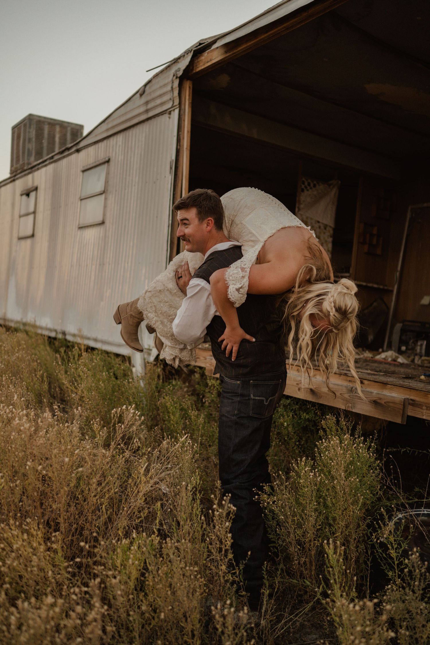 Playful wedding photo of couple at abandoned trailer during golden hour.