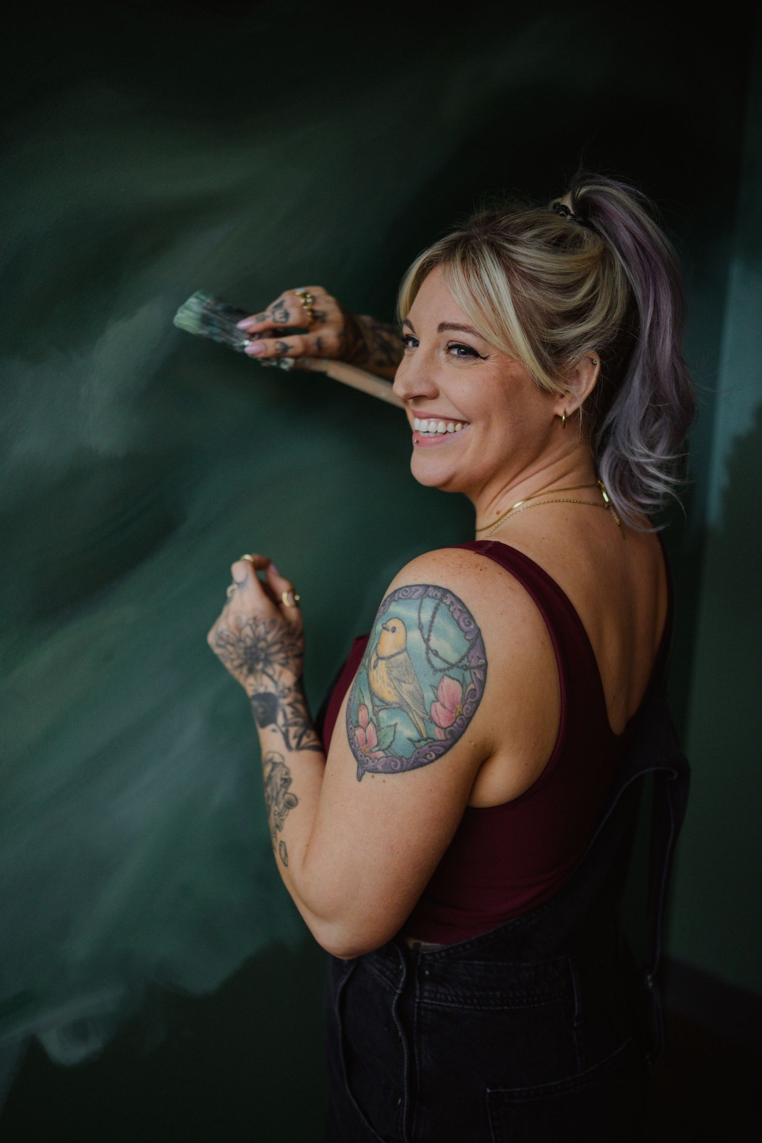 An artist with colorful tattoos works on painting against a dark green background.
