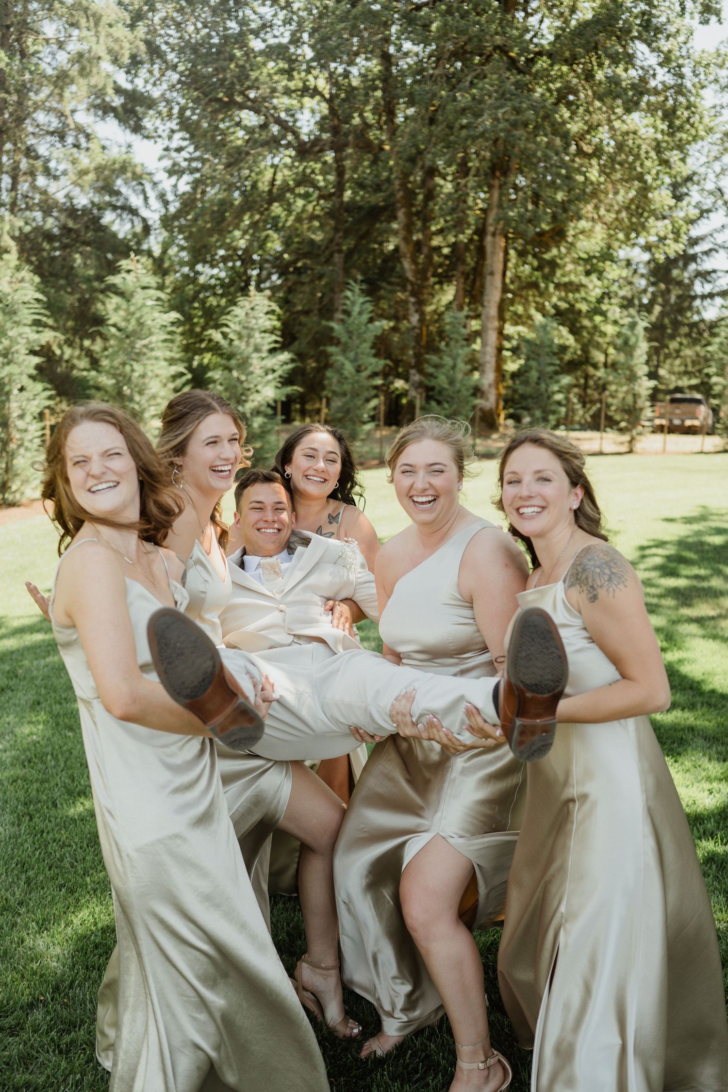 Wedding party in champagne silk dresses playfully lifting someone up on a green lawn.