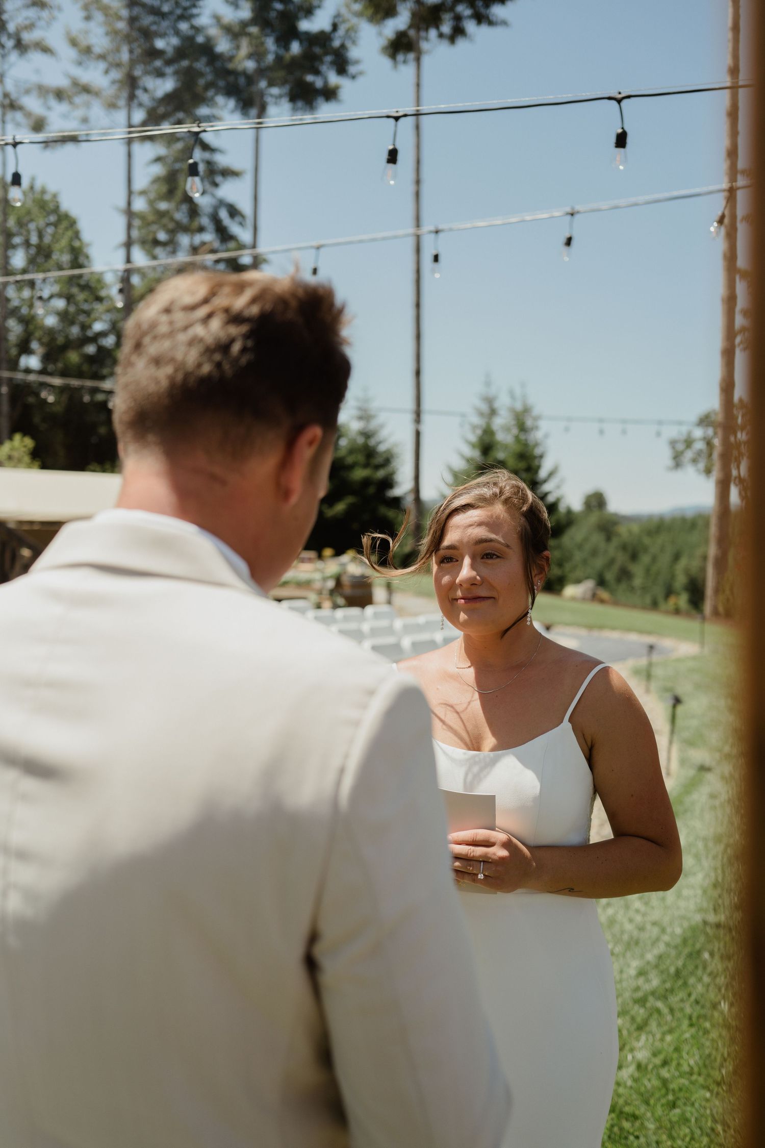 A couple exchanging vows during an outdoor wedding ceremony with string lights visible above.