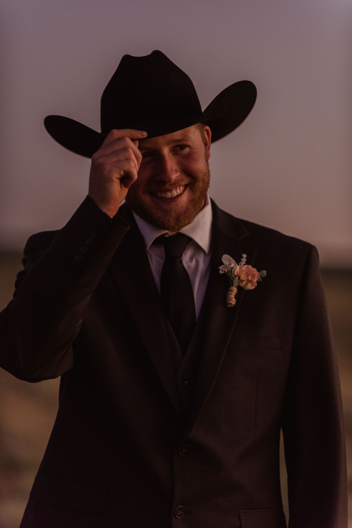 A western-styled groom in black suit and cowboy hat smiles while adjusting his hat.
