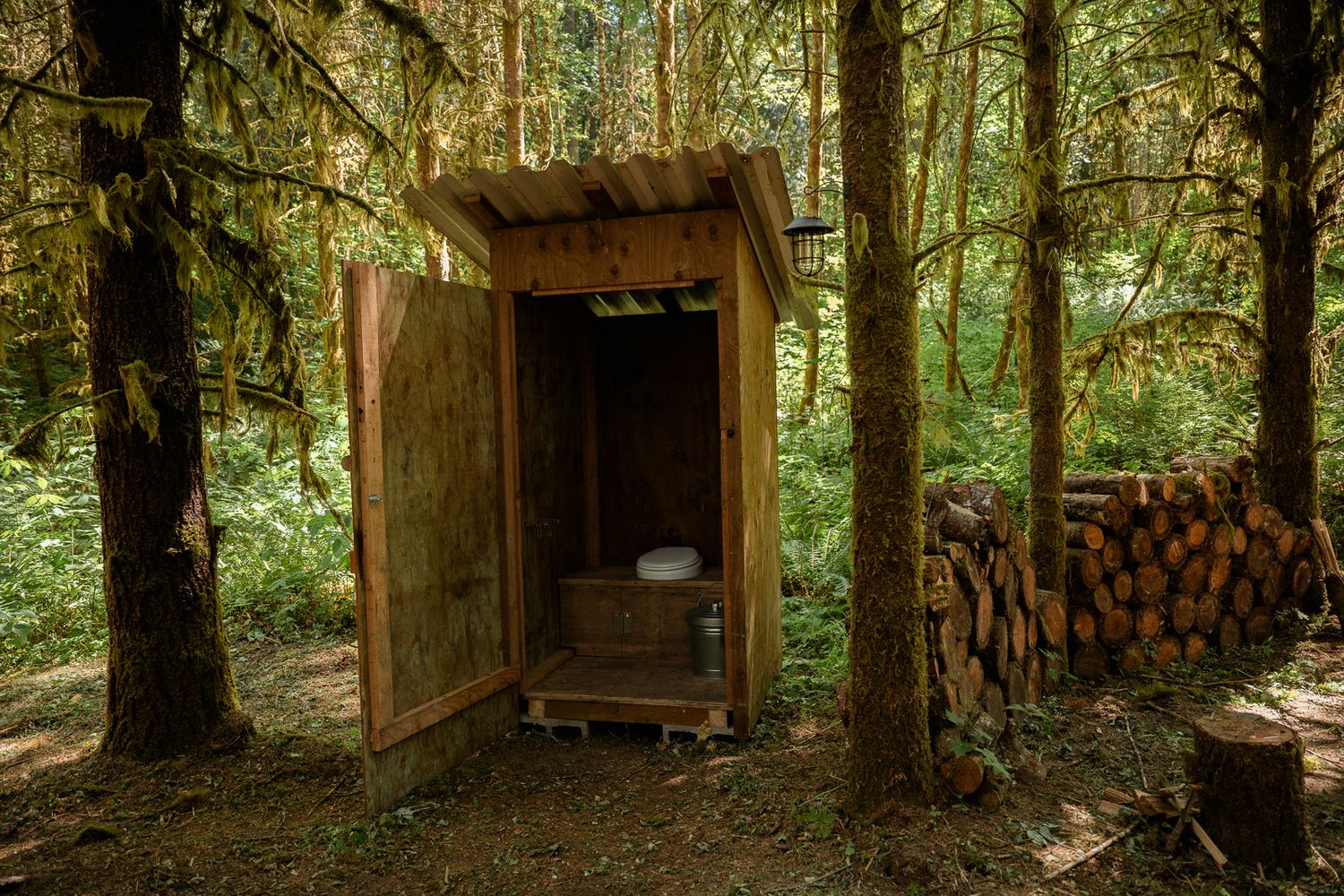 Rustic wooden outhouse facility in forest clearing surrounded by stacked firewood.