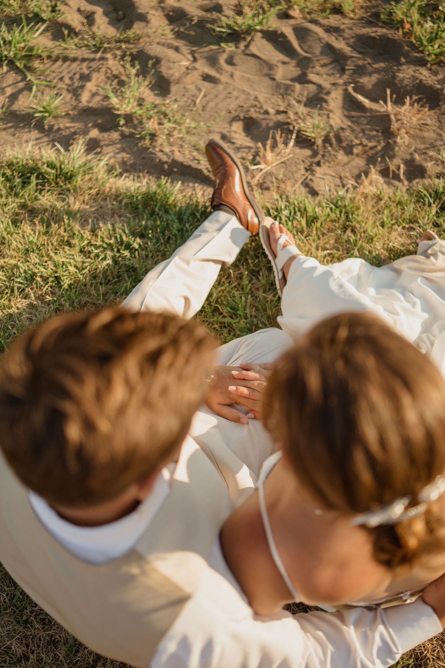 A couple relaxes on grass wearing white outfits during a sunny outdoor photoshoot.