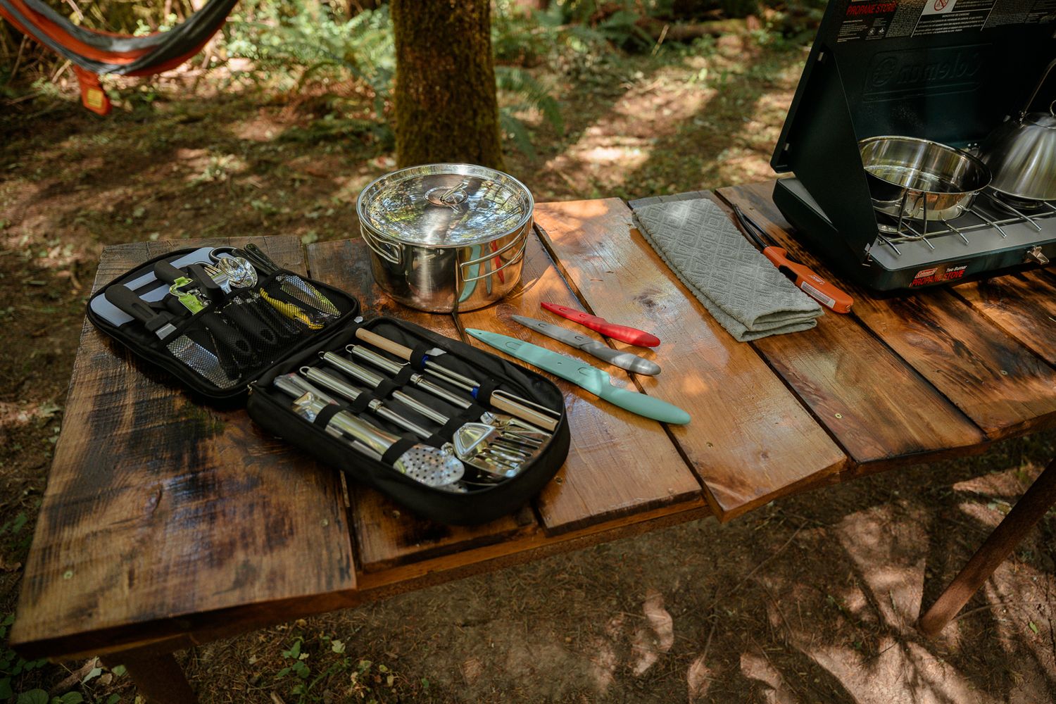 Camping cooking supplies and utensils laid out on wooden surface in forest setting.