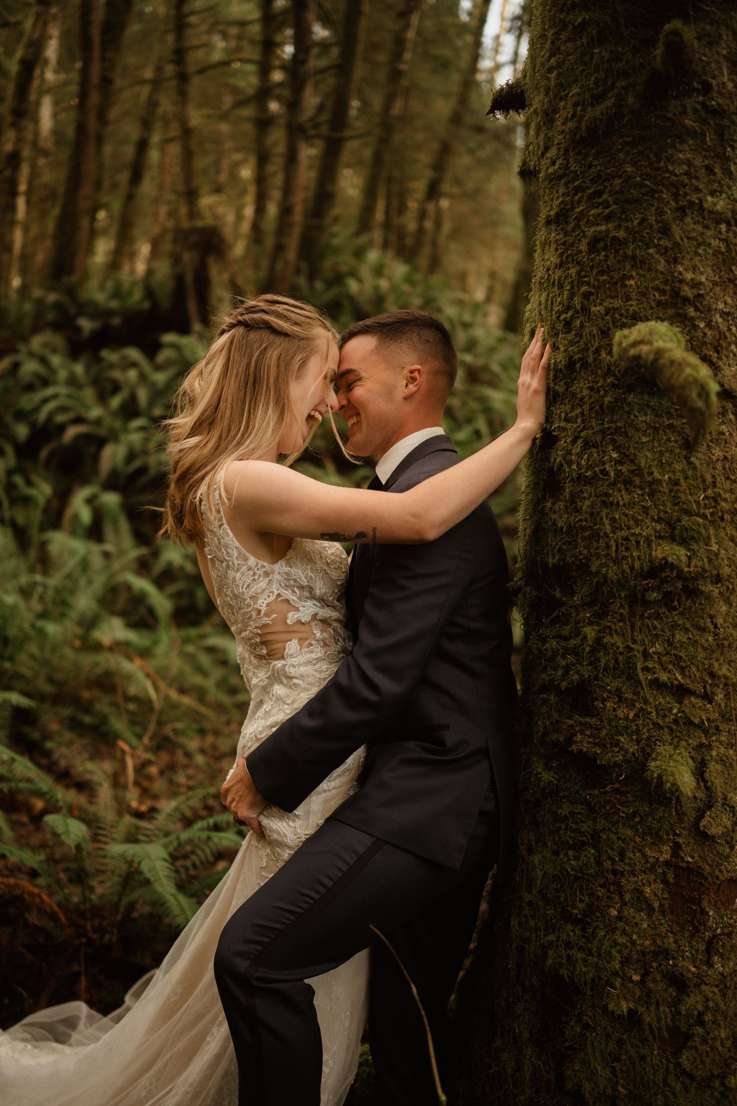 A romantic wedding portrait of a couple embracing against a mossy tree trunk in a lush forest setting.