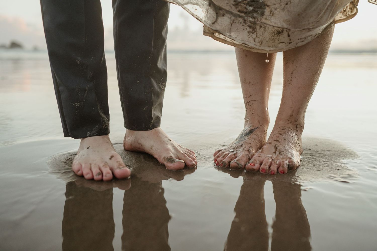 Bare feet of a couple standing in wet sand at the beach during sunset.