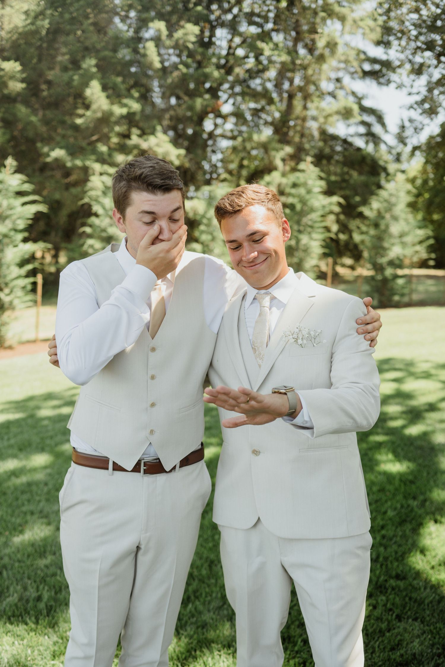 Two people in white suits share a candid moment of laughter on a sunny lawn.