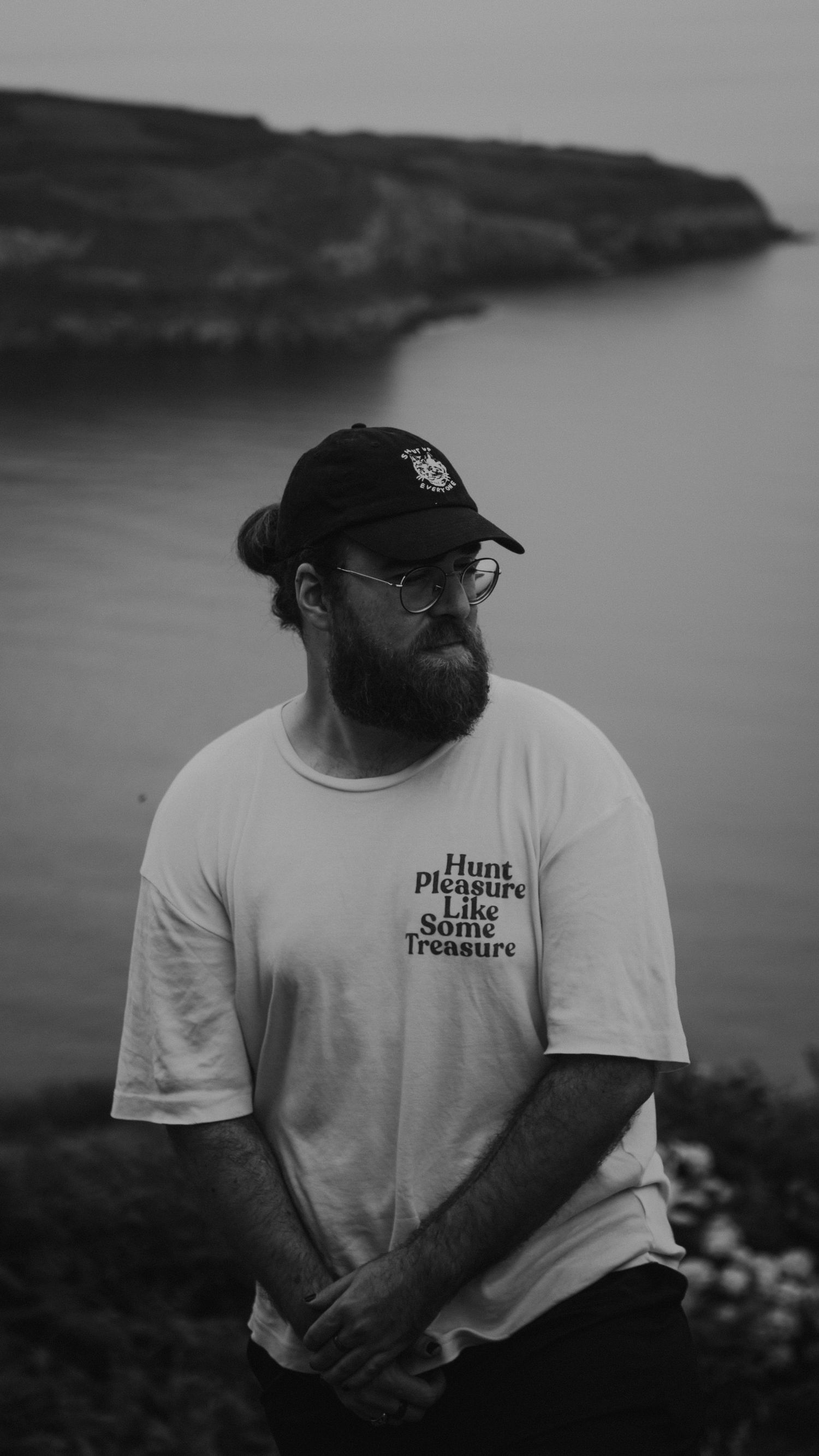A photographer in casual attire stands against a coastal landscape in black and white.