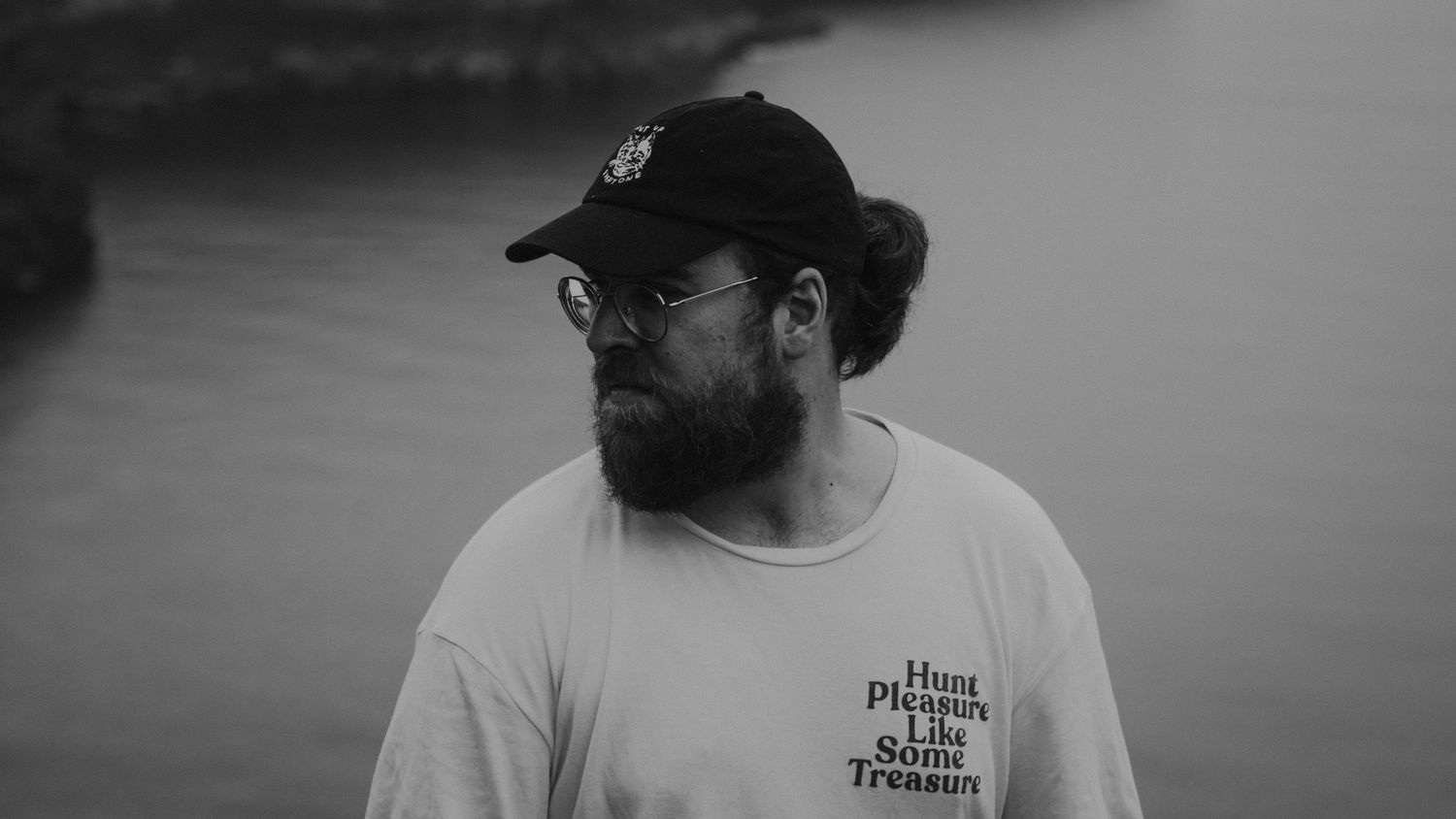 A casual portrait of a photographer against a serene waterfront backdrop in monochrome.