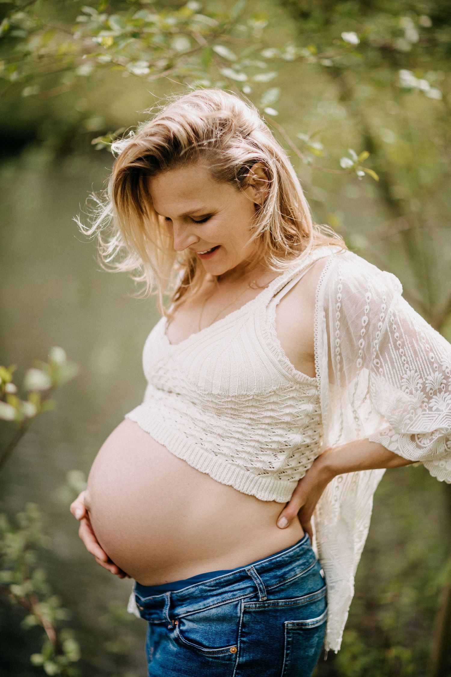 Eine werdende Mutter in einem weißen Oberteil steht draußen und berührt ihren schwangeren Bauch.