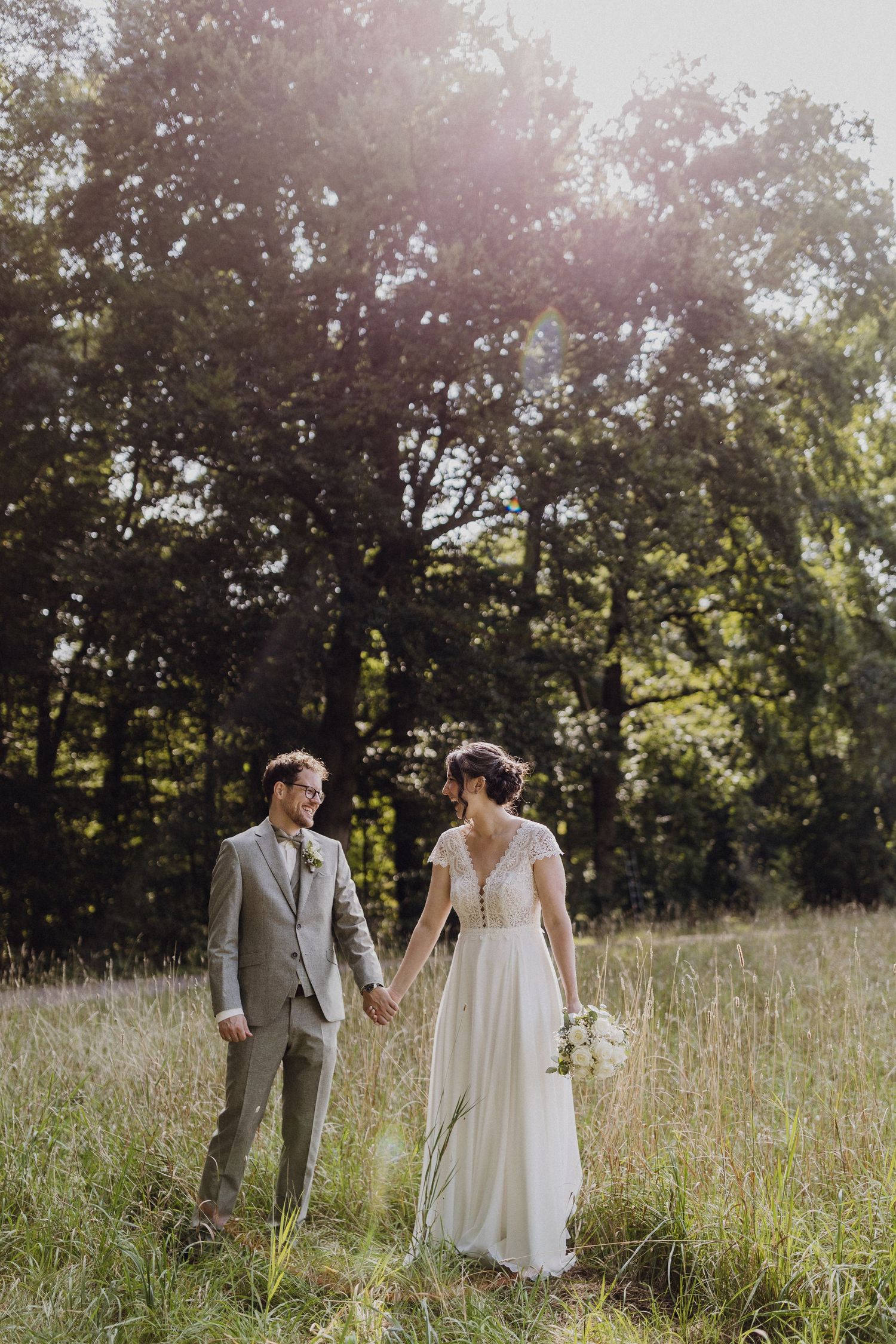 Ein Hochzeitspaar geht Hand in Hand durch eine sonnenbeschienene Wiese mit Bäumen im Hintergrund.