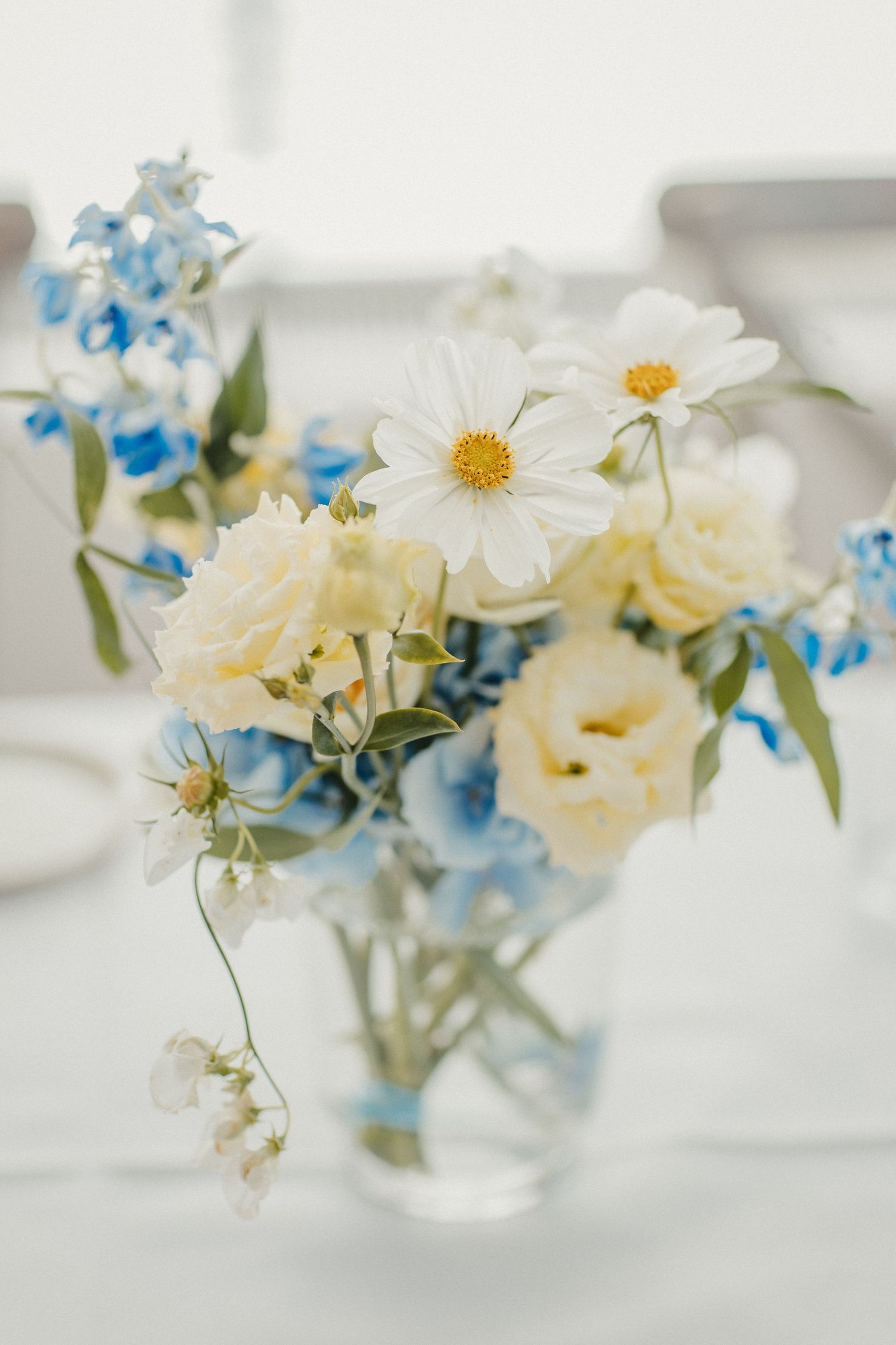 Zartes Blumenarrangement mit weißen Gänseblümchen, cremefarbenen Rosen und blauem Delphinium in einer klaren Glasvase.