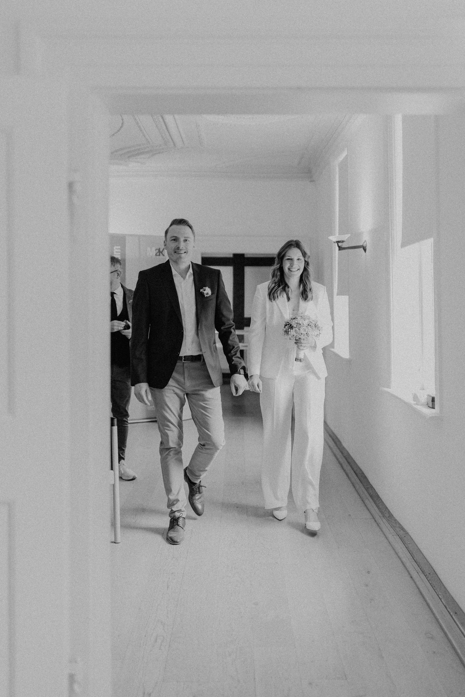 Schwarz-Weiß-Foto eines Paares, das in lässiger Hochzeitskleidung den Flur entlang geht.