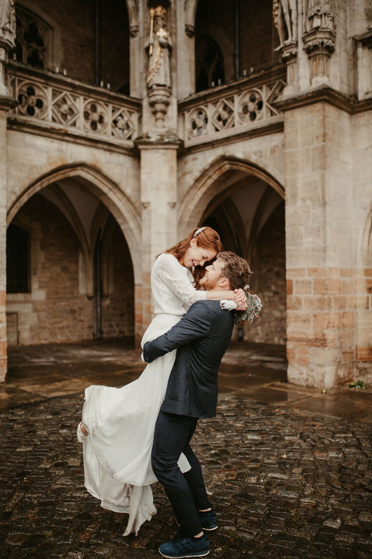 Romantisches Hochzeitspaar umarmt im gotischen Kathedralenhof mit Steinbögen.