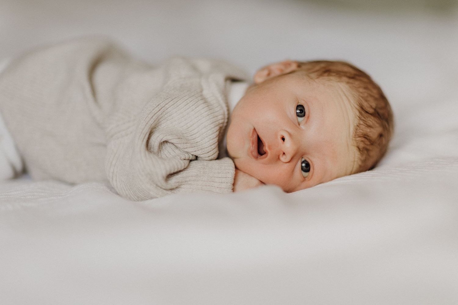 Baby in weichem beigem Pullover liegt auf weißer Bettwäsche.