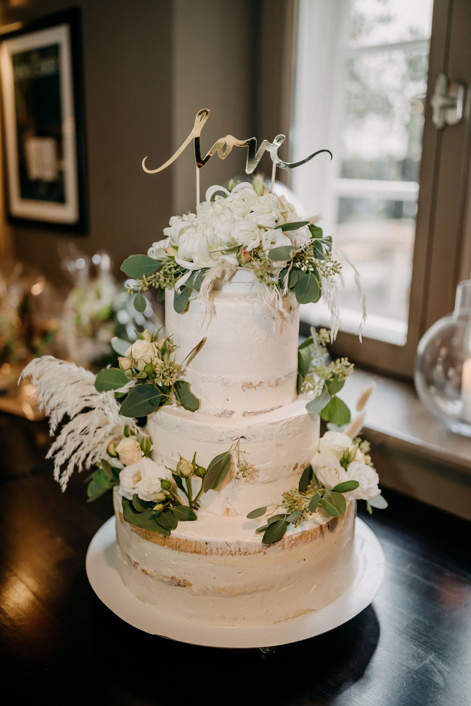 Dreistufige weiße Hochzeitstorte, verziert mit Grün und goldenen Akzenten, gekrönt mit einem Liebeszeichen.