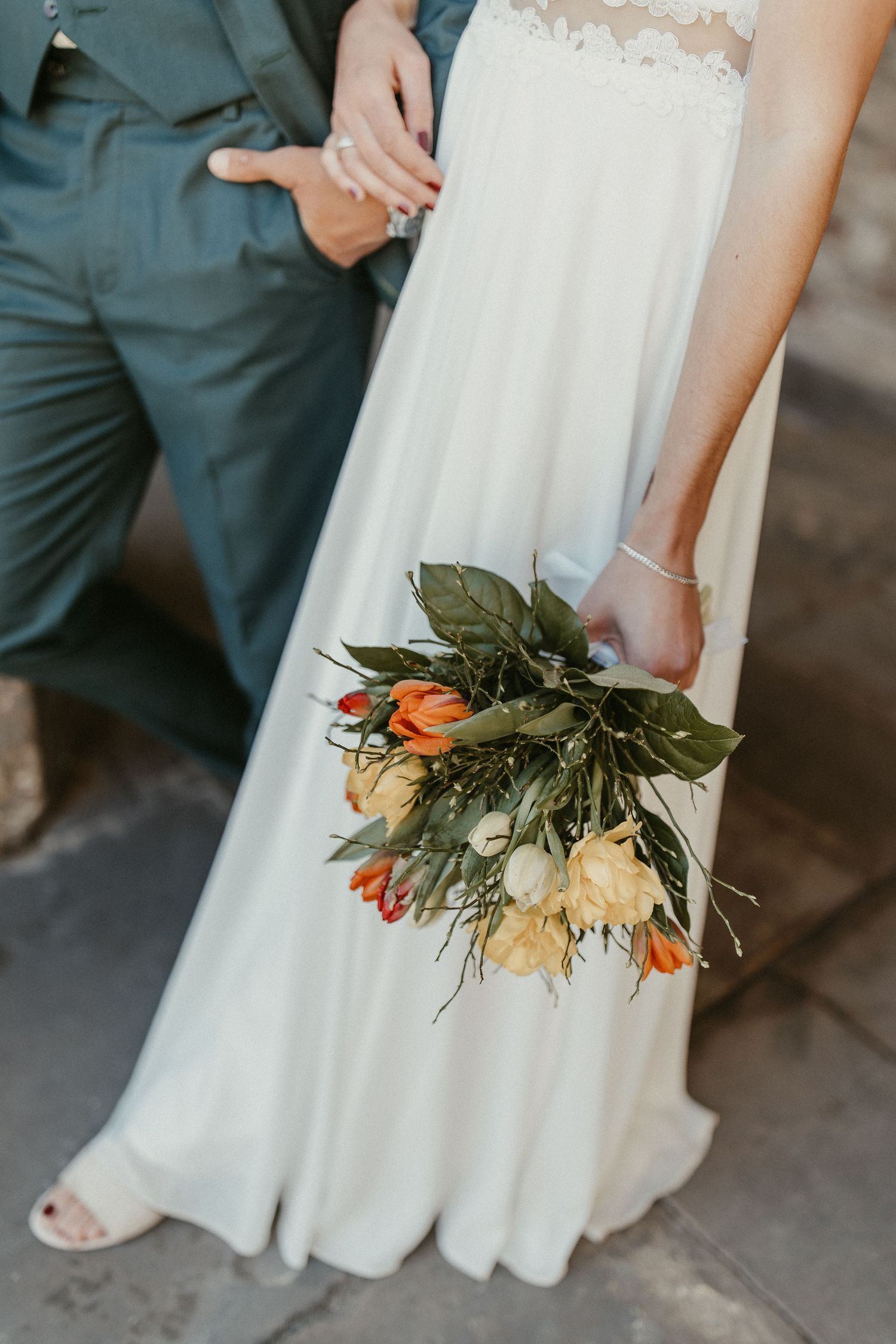 Eine Nahaufnahme eines Brautstraußes mit orangefarbenen und cremefarbenen Blumen vor einem weißen Hochzeitskleid.