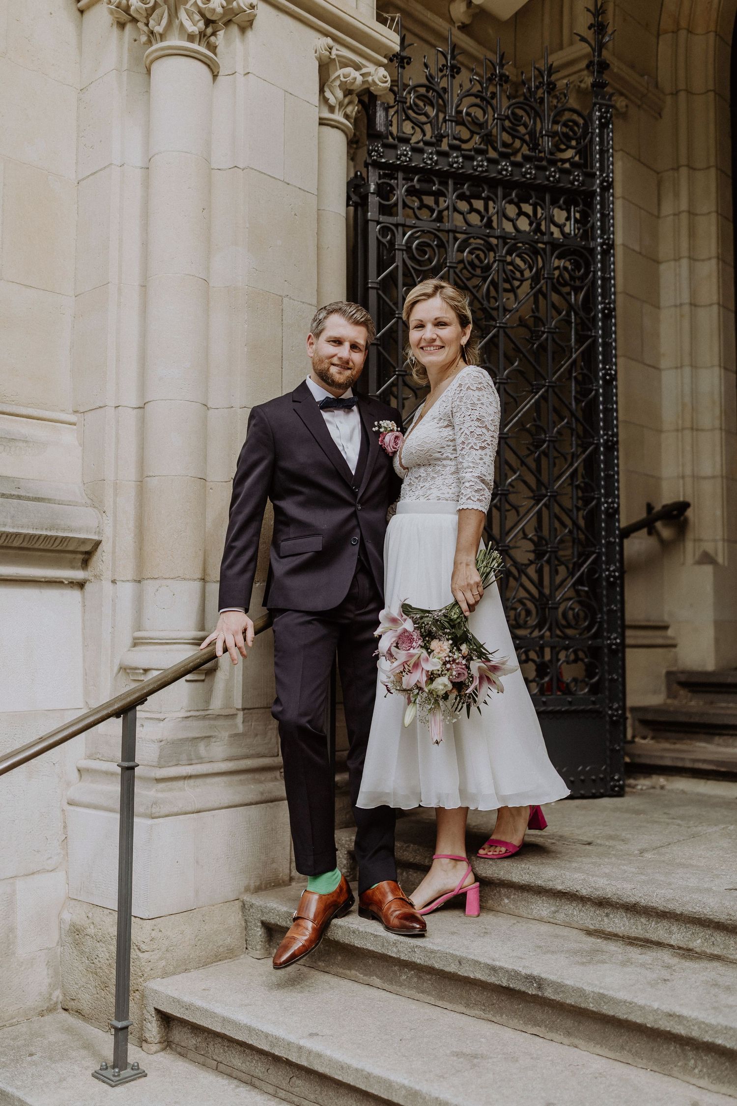Elegantes Paar auf Kirchenstufen mit kunstvoller Eisentür und Vintage-inspirierter Hochzeitskleidung.