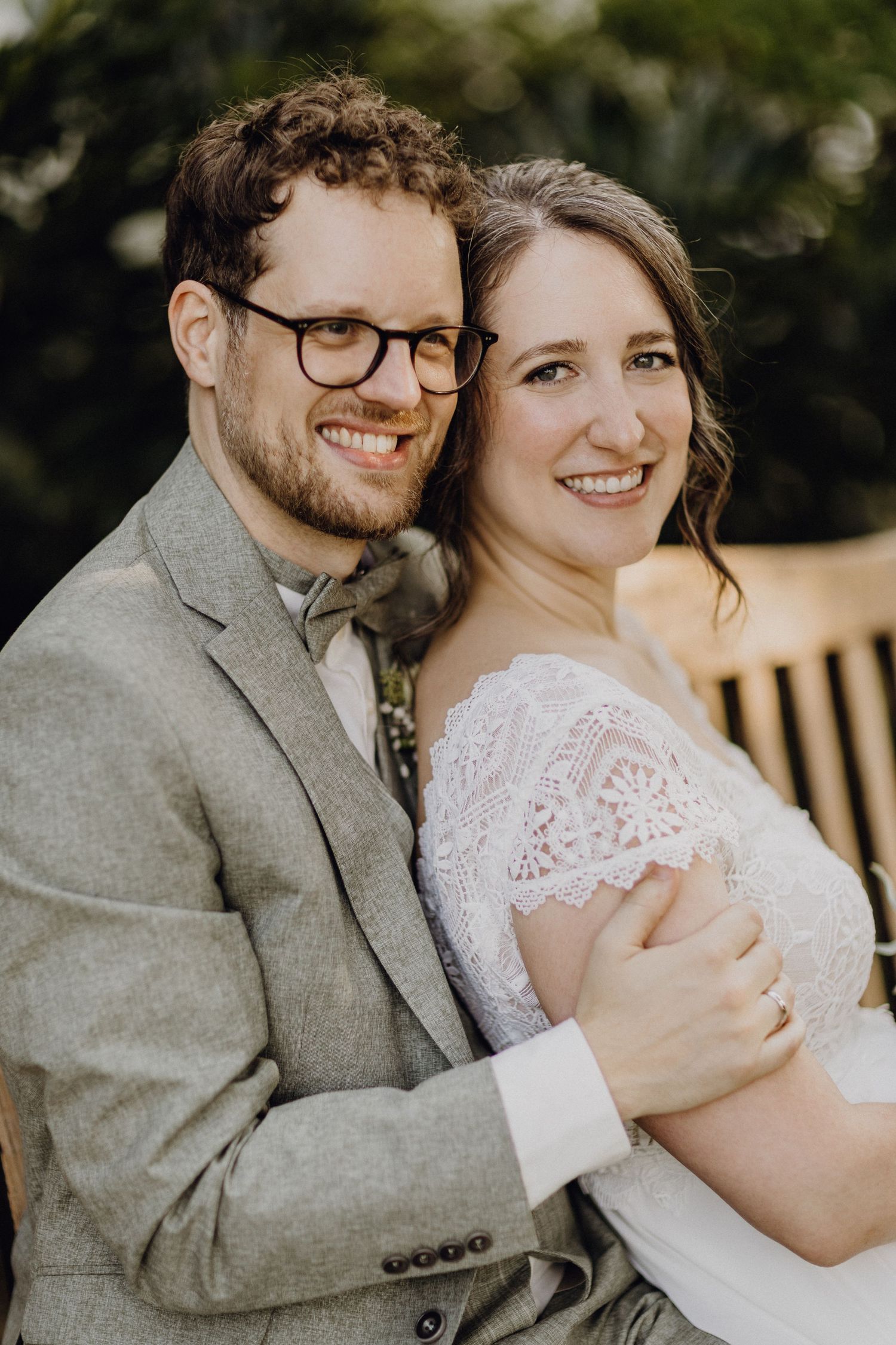 Ein romantisches Hochzeitsporträt eines Paares in grauem Anzug und Spitzenkleid, das draußen zusammen lächelt.